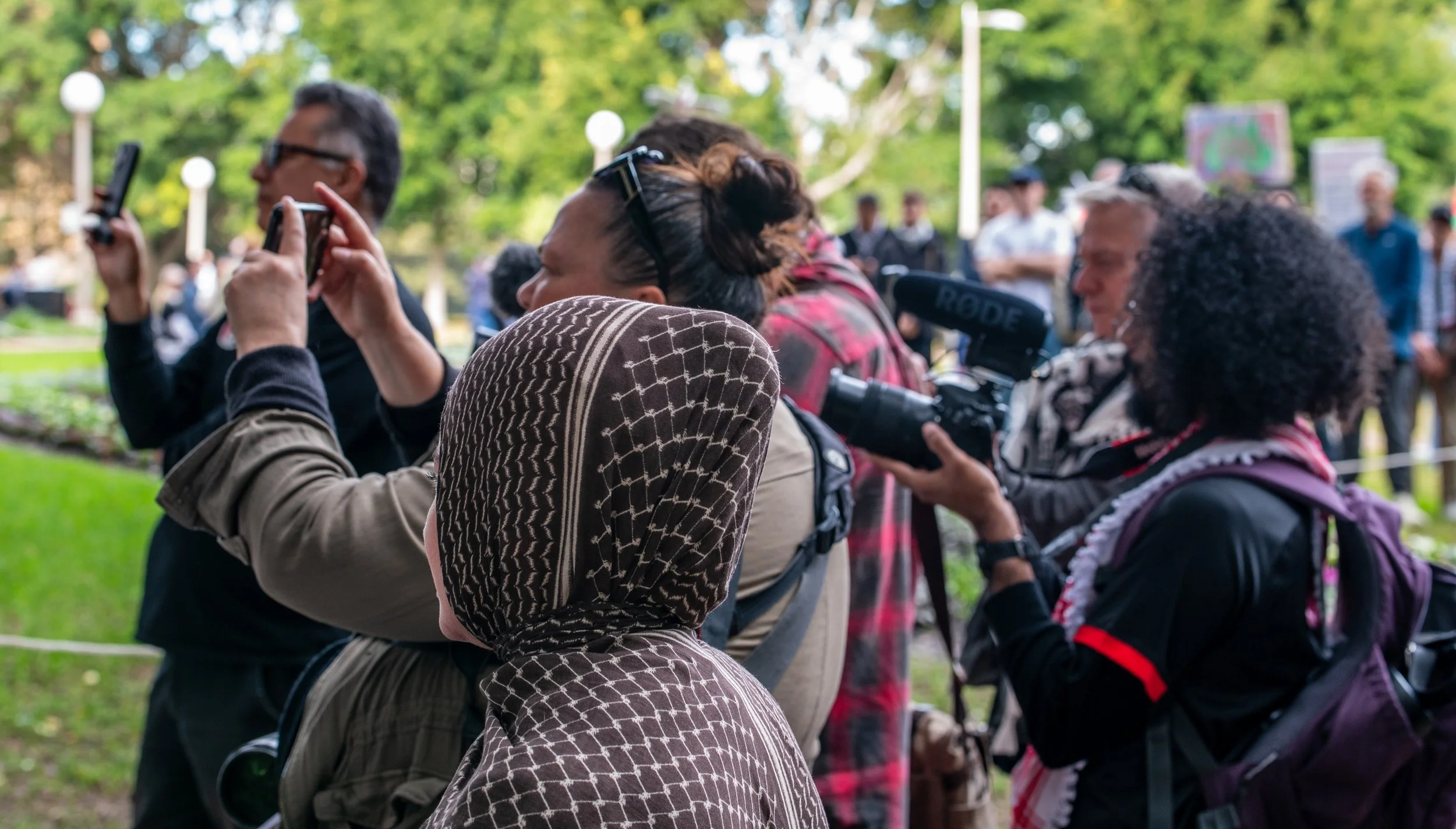 People gathered outdoors, some taking photos or videos with smartphones and cameras, at a park with green trees in the background.