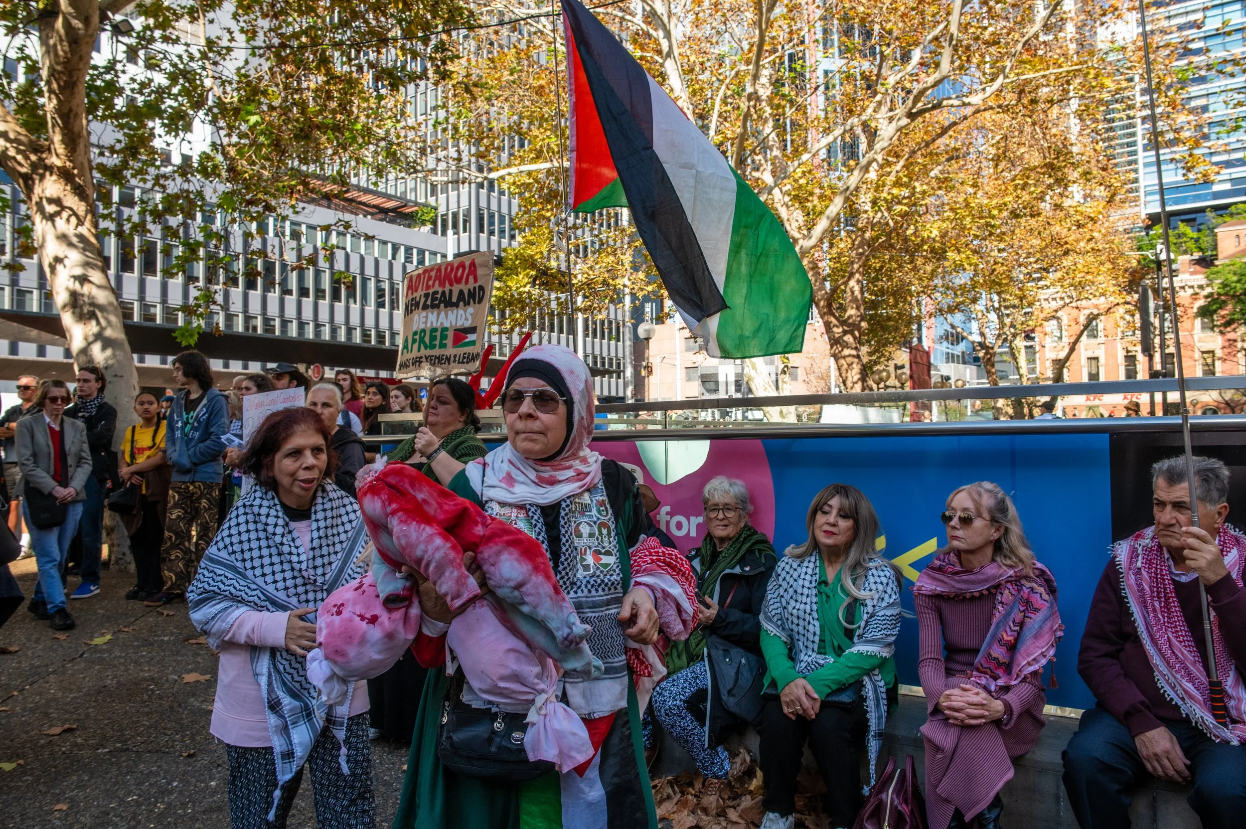 A group of protesters gathered outdoors, some wearing keffiyeh scarves, with a Palestinian flag flying, and holding signs supporting Palestine and demanding independence, against a backdrop of trees and modern buildings.