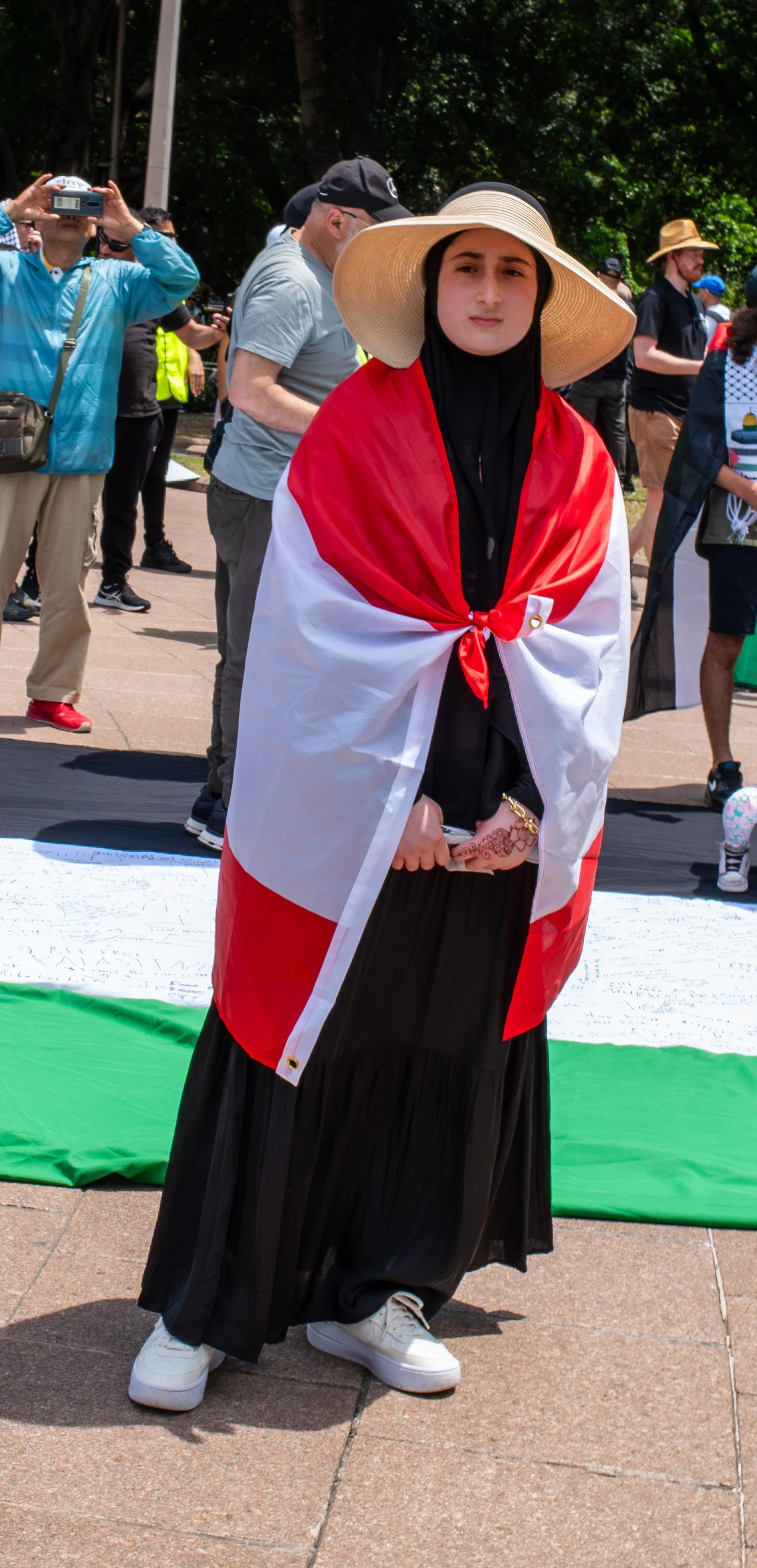 Woman wearing a large straw hat and black clothing, wrapped in a flag with red and white colors, standing outdoors during a gathering or protest.