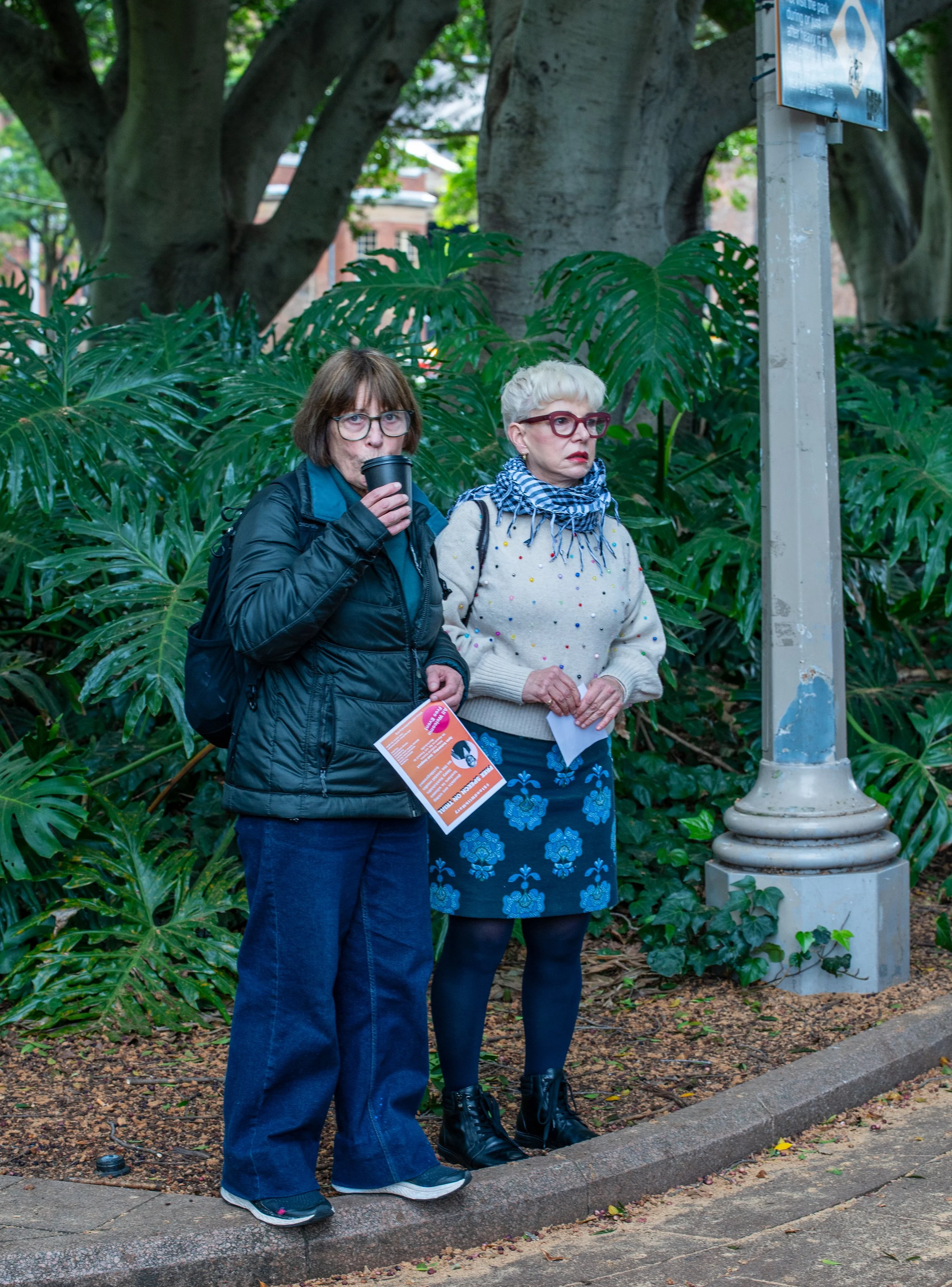 Two women standing near large green leafy plants, one drinking from a cup, the other holding papers, in a park setting with large trees and a street sign.