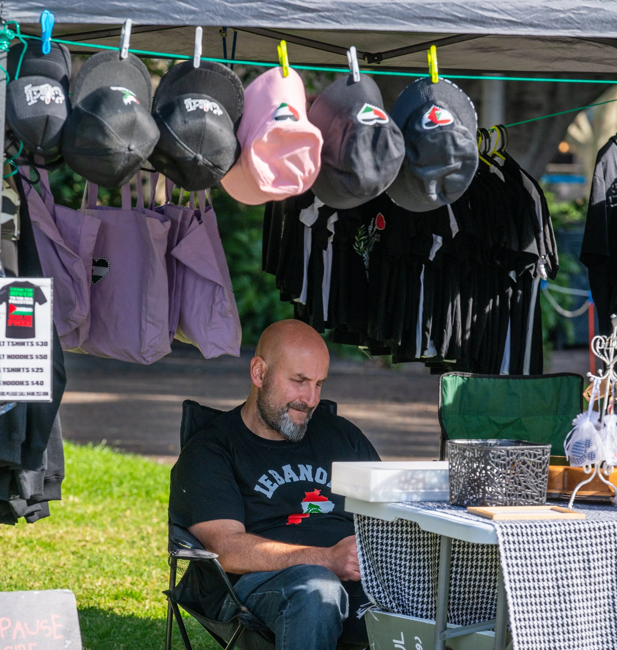 Man sitting at a booth selling hats, bags, and clothing outdoors, under a tent, with various caps hanging above him.