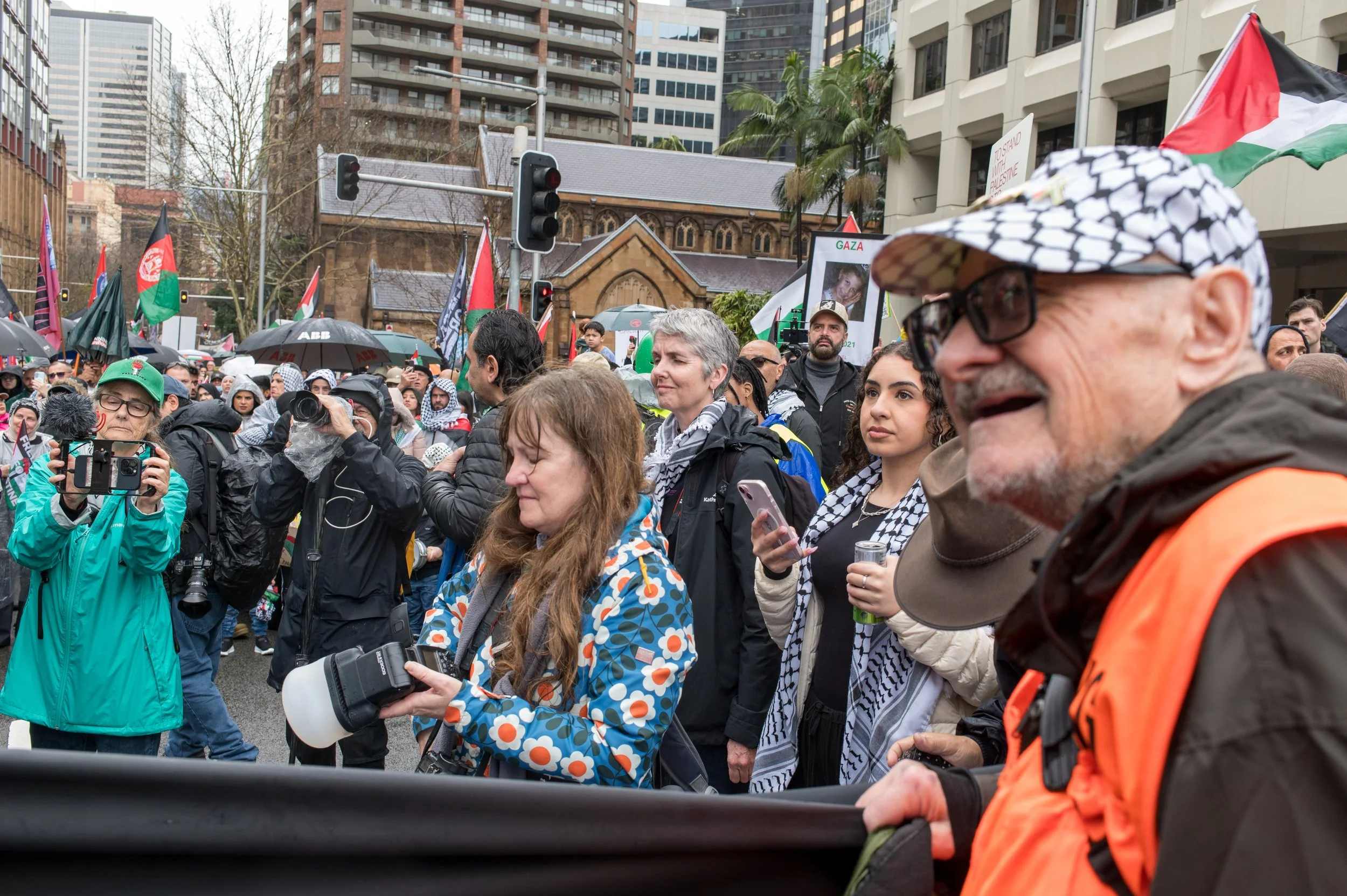 Crowd of people protesting outdoors under cloudy sky, holding flags and signs, some wearing Palestinian keffiyeh scarves, in an urban area with buildings and traffic lights.