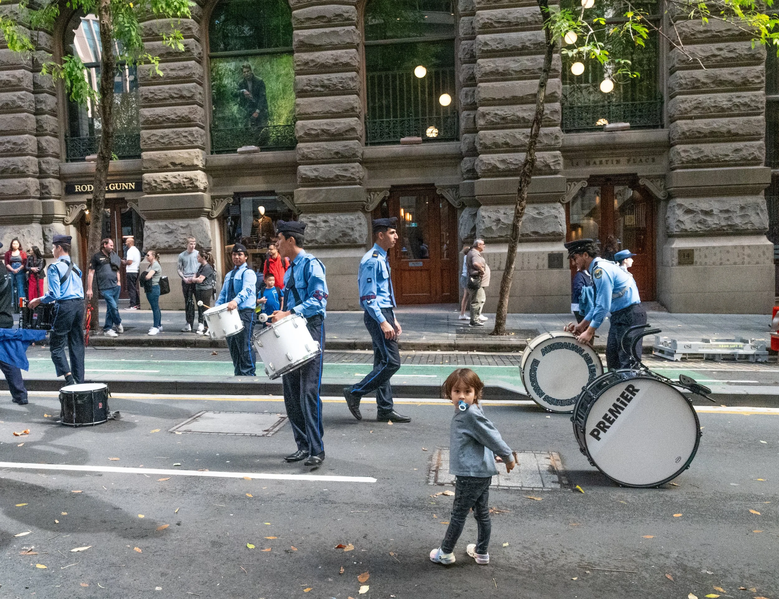 A street scene in a city with a drum band made up of young men dressed in blue uniforms. A little girl with a pacifier stands in the foreground, looking at the camera. Pedestrians walk along the sidewalk, and the background features a stone building with large windows and trees.