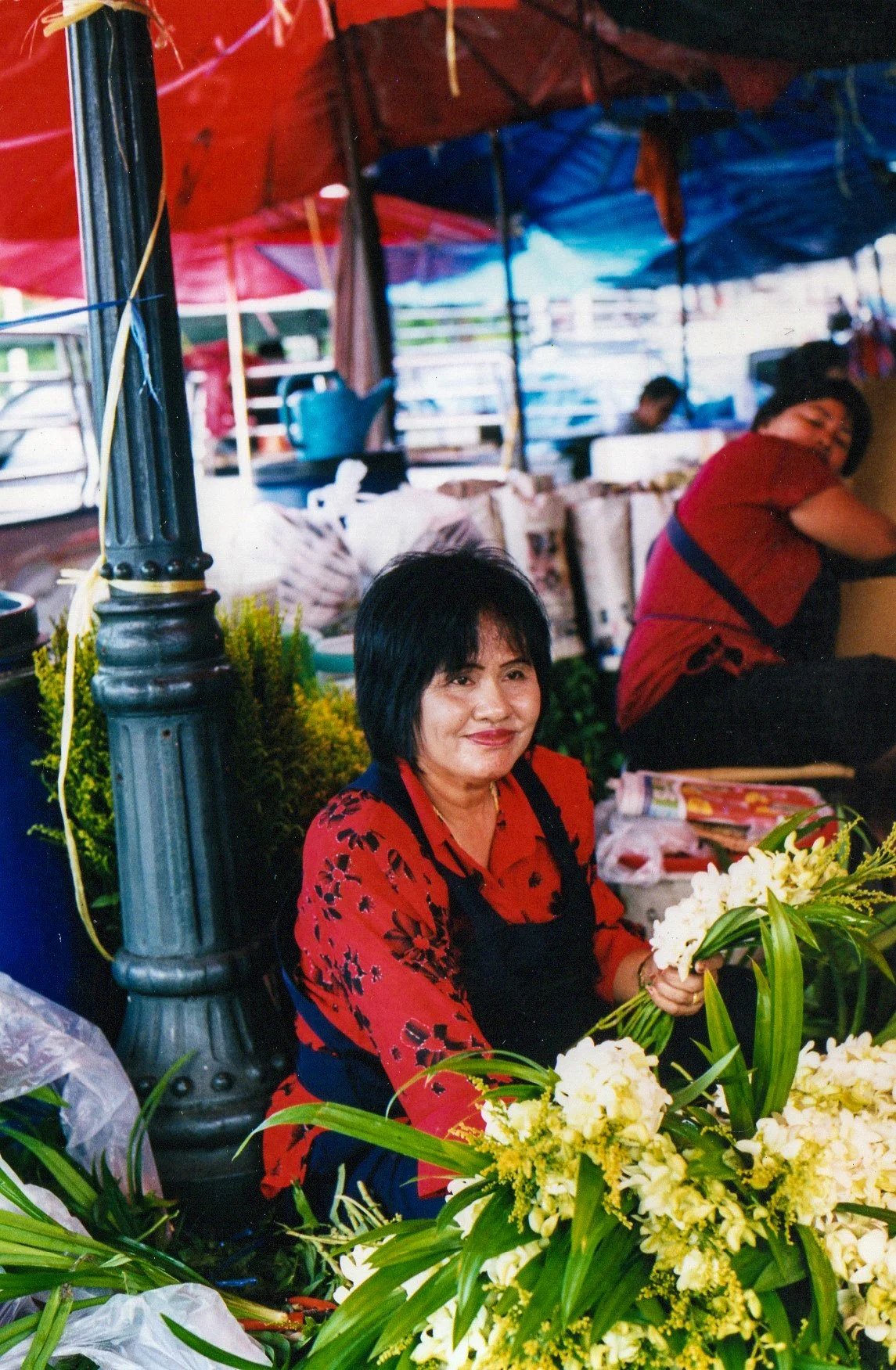 A woman with short dark hair, wearing a red floral shirt and black apron, sitting in a market stall surrounded by yellow and white flowers, holding a bouquet of flowers and smiling.