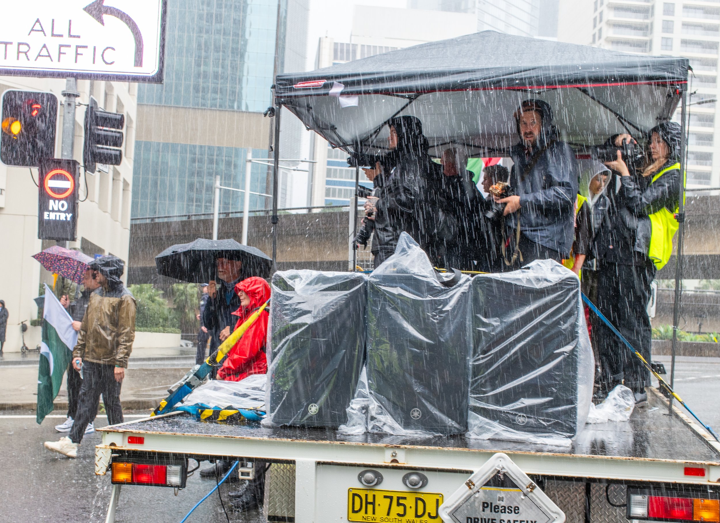 A group of people on a truck float during a rainy day, some holding umbrellas and a person carrying a flag, with tall buildings and traffic signals in the background.