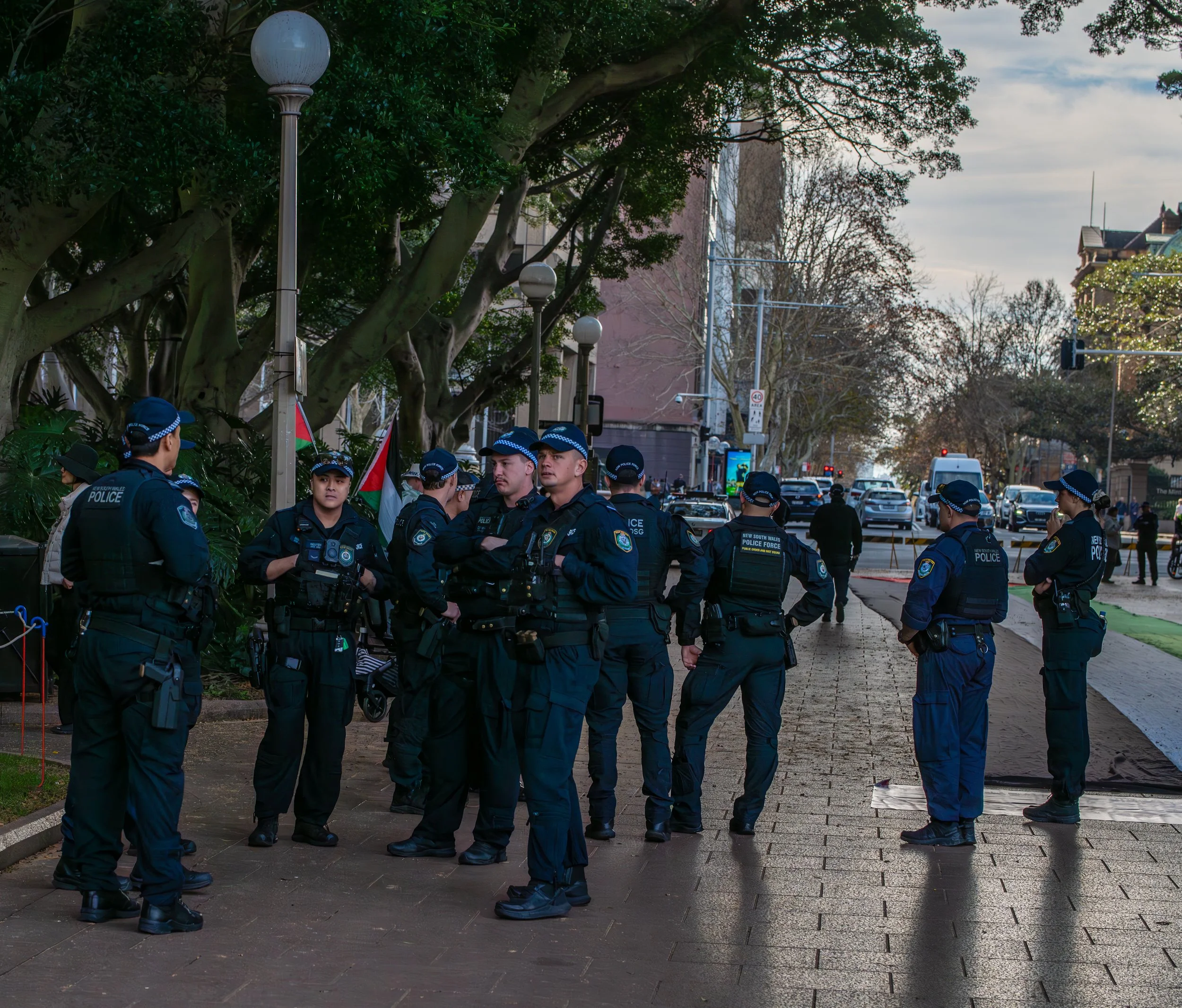Group of police officers standing and talking on a sidewalk in an urban area during daytime, with several cars and people in the background.