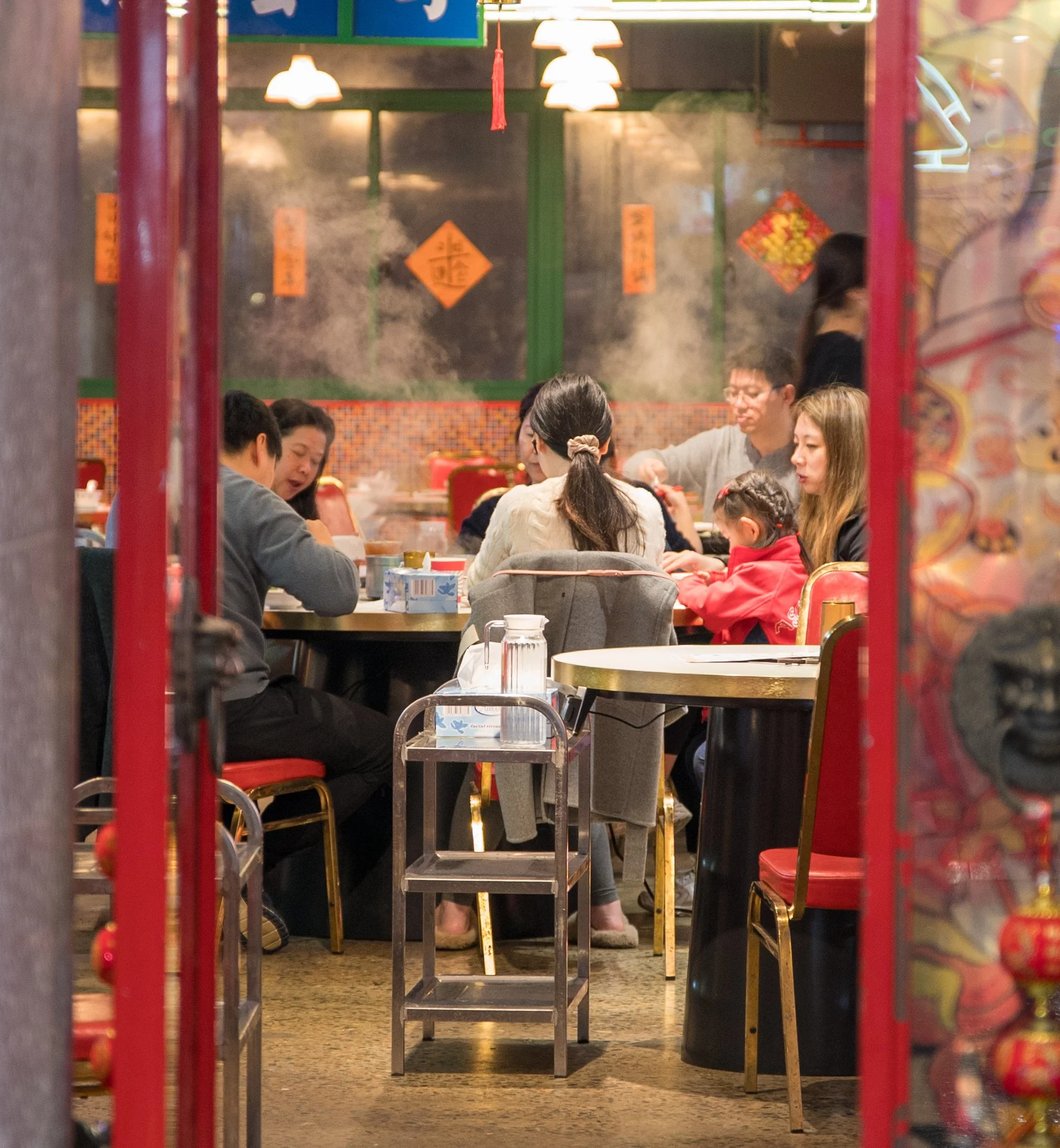 A group of people dining together in a Chinese restaurant with red and gold decorations, including lanterns and wall hangings, seen through a doorway.