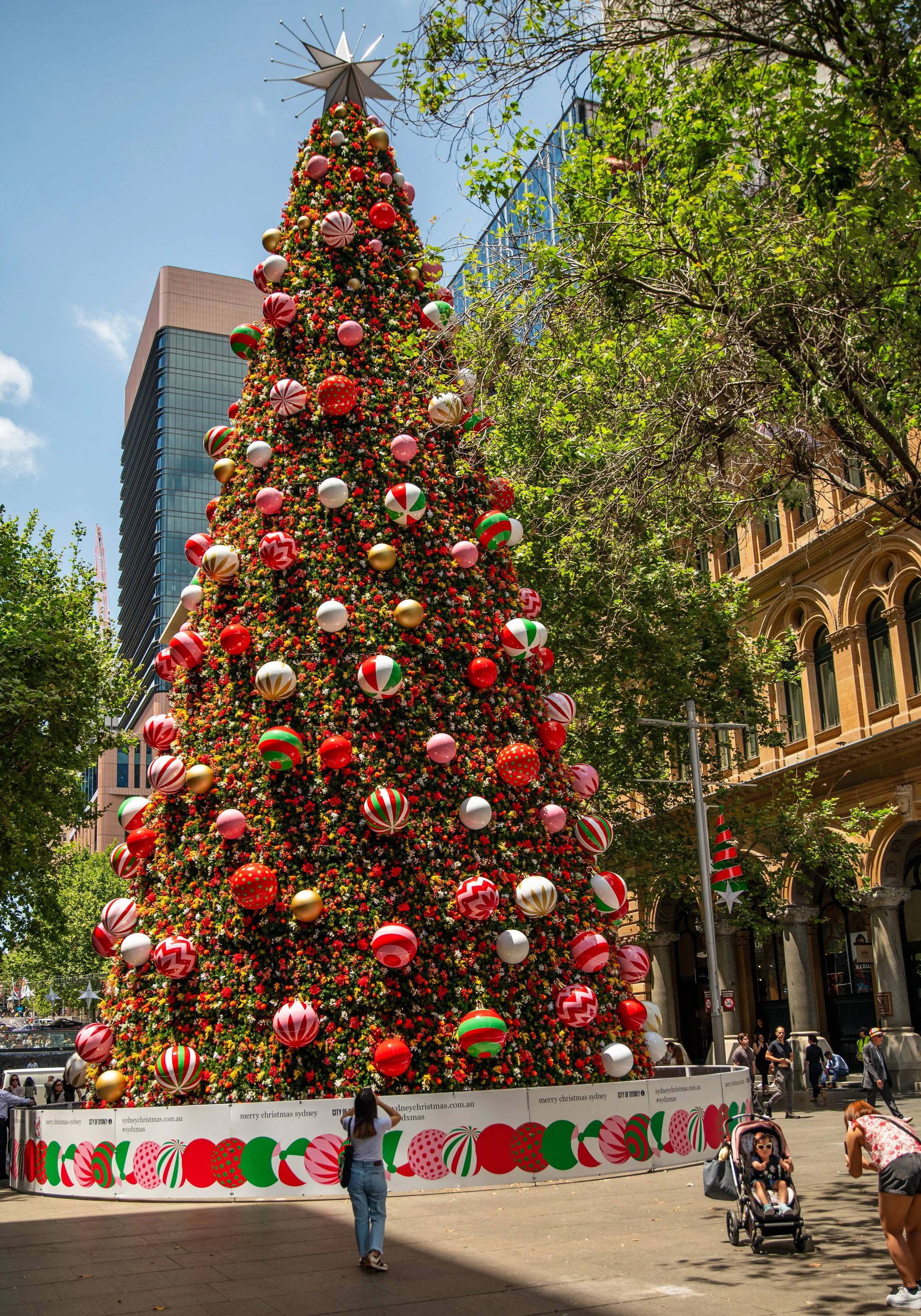 A tall Christmas tree decorated with large red, white, pink, green, and gold ornaments, topped with a silver star, in an urban city square surrounded by trees and historic buildings, with people walking and a woman taking a photo.