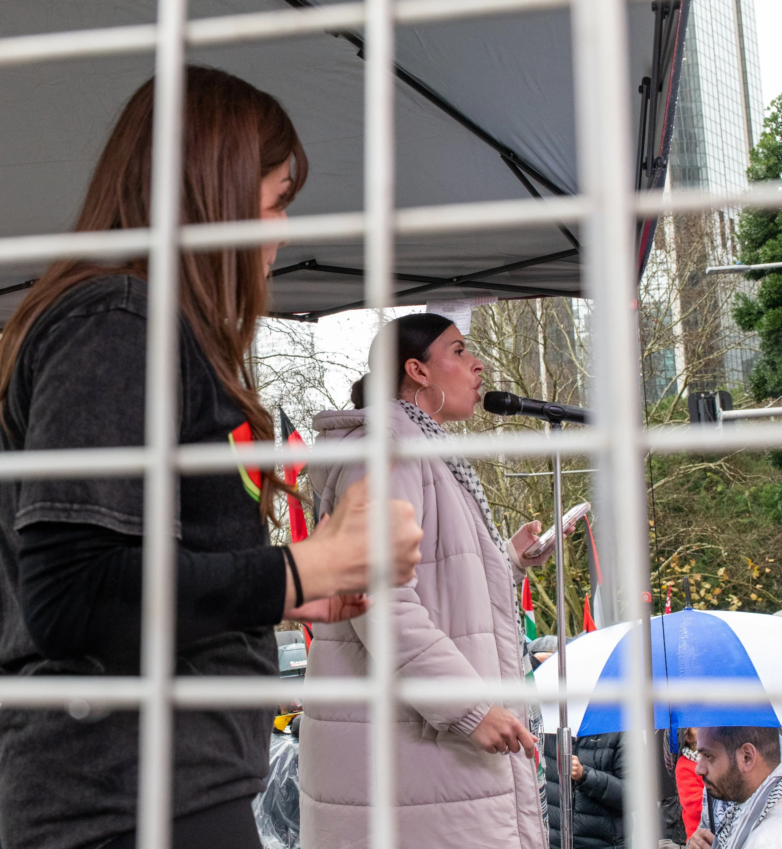 Women speaking at a protest or rally, seen through a metal fence with trees and buildings in the background.