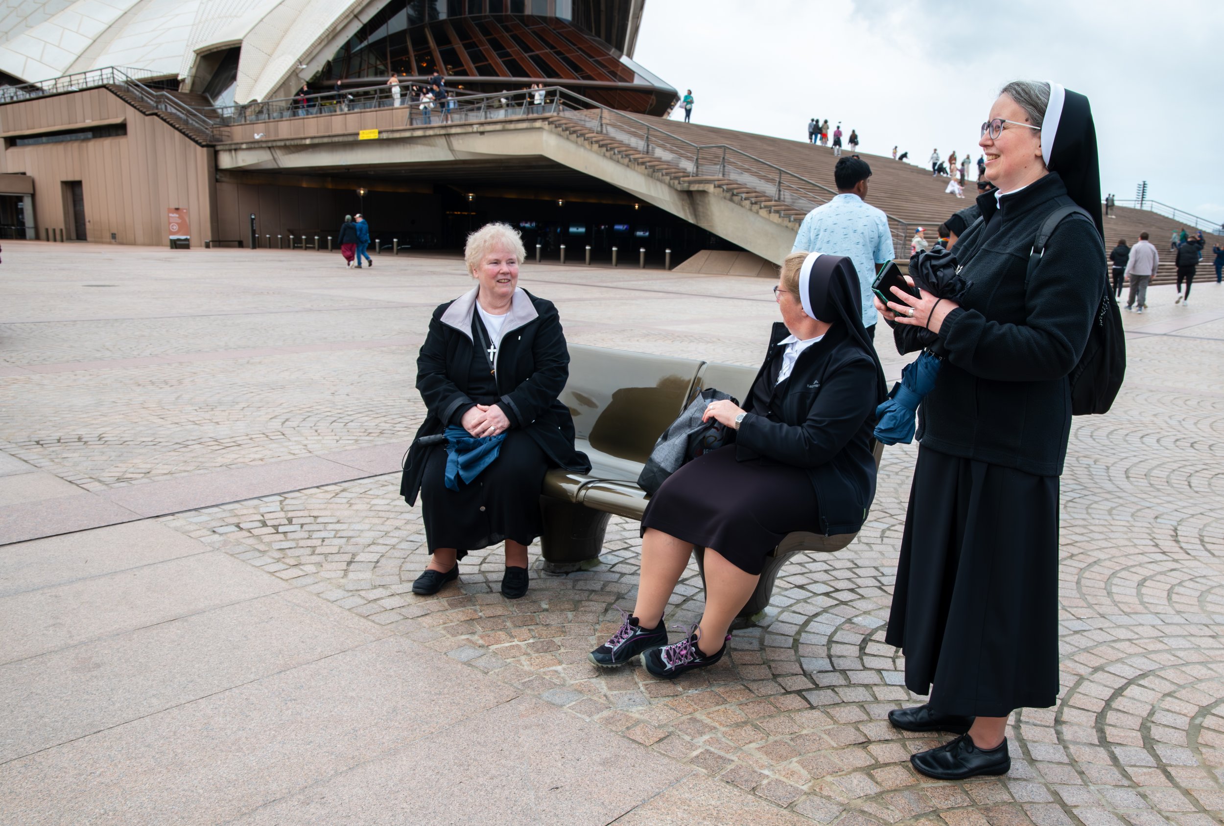 Three women, two dressed as nuns, are having a conversation on a bench outside the Sydney Opera House in Australia. One woman is sitting on the bench, smiling, while the other two stand nearby, one of whom is holding a phone. The background shows the iconic sail-like architecture of the Sydney Opera House and a few other people walking or standing around.