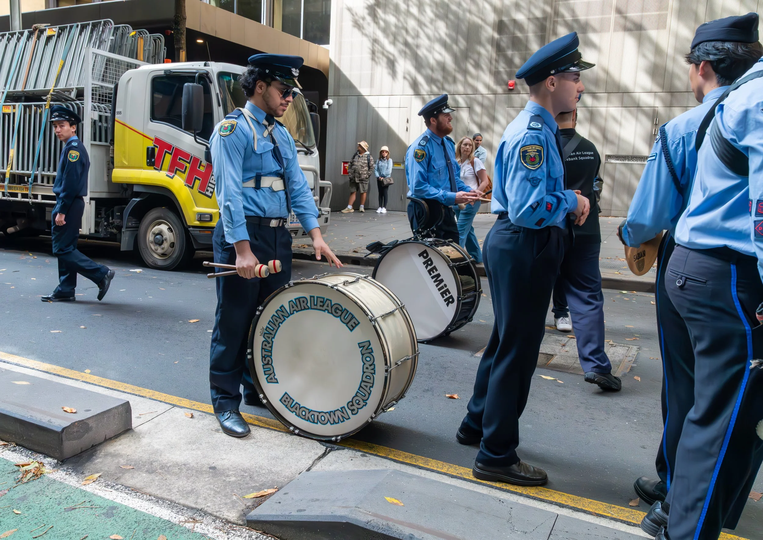 Group of uniformed musicians from the Australasia Air League Blacktown Squadron standing on a city street, some holding drums and percussion instruments, with a truck and onlookers in the background.