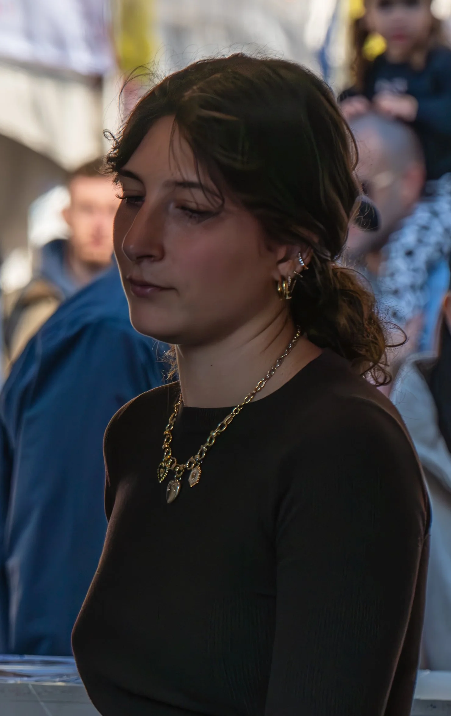 A young woman with dark hair and multiple earrings, wearing a black top and layered gold necklaces, standing with her eyes closed or looking down in a crowded setting.