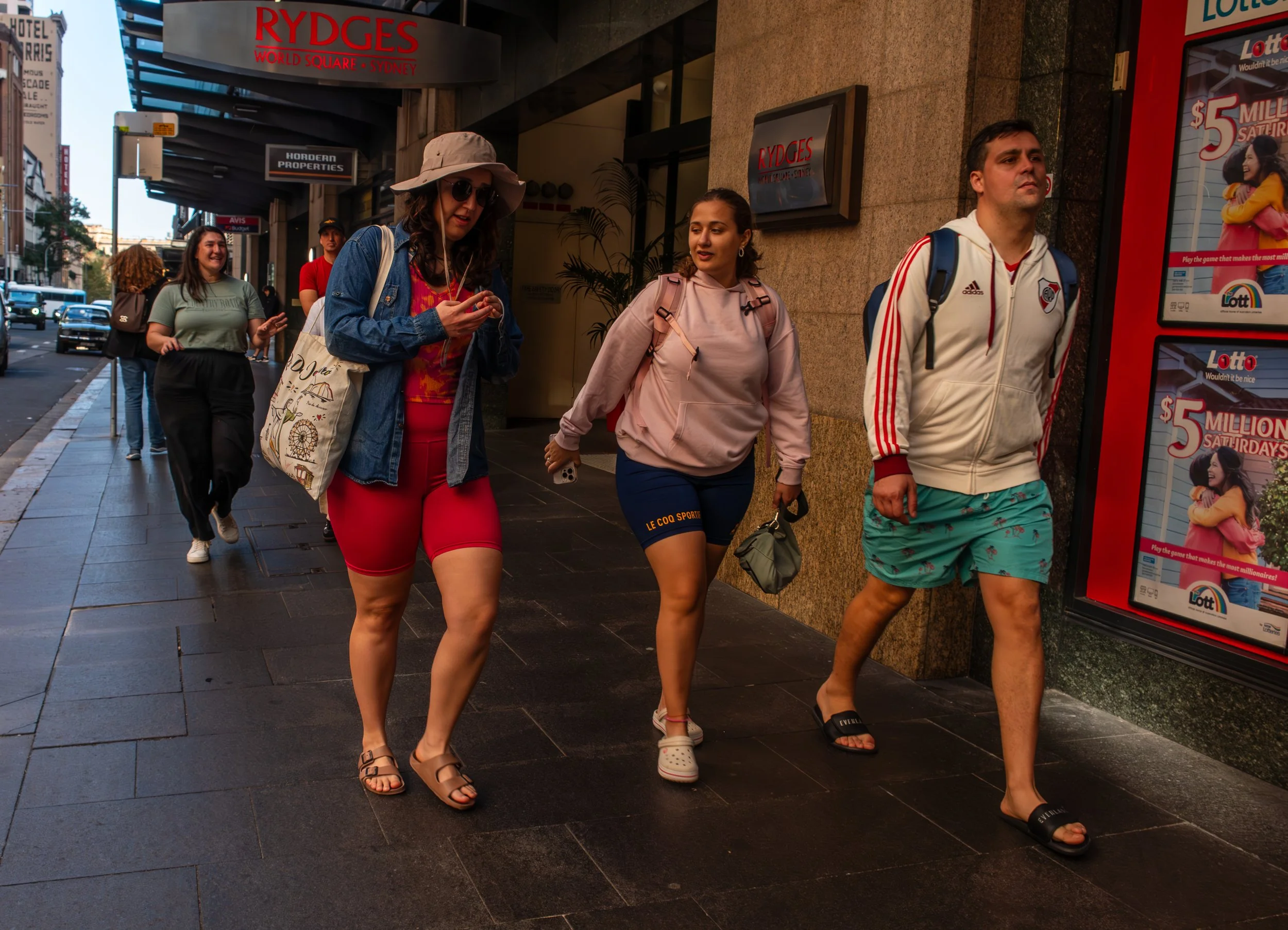 A group of five people walking on a city sidewalk, two women in the foreground wearing casual clothes and backpacks, a man in shorts and a hoodie, and two women in the background, one with curly hair and the other with straight hair, all smiling or focused, with buildings and advertisements along the street.