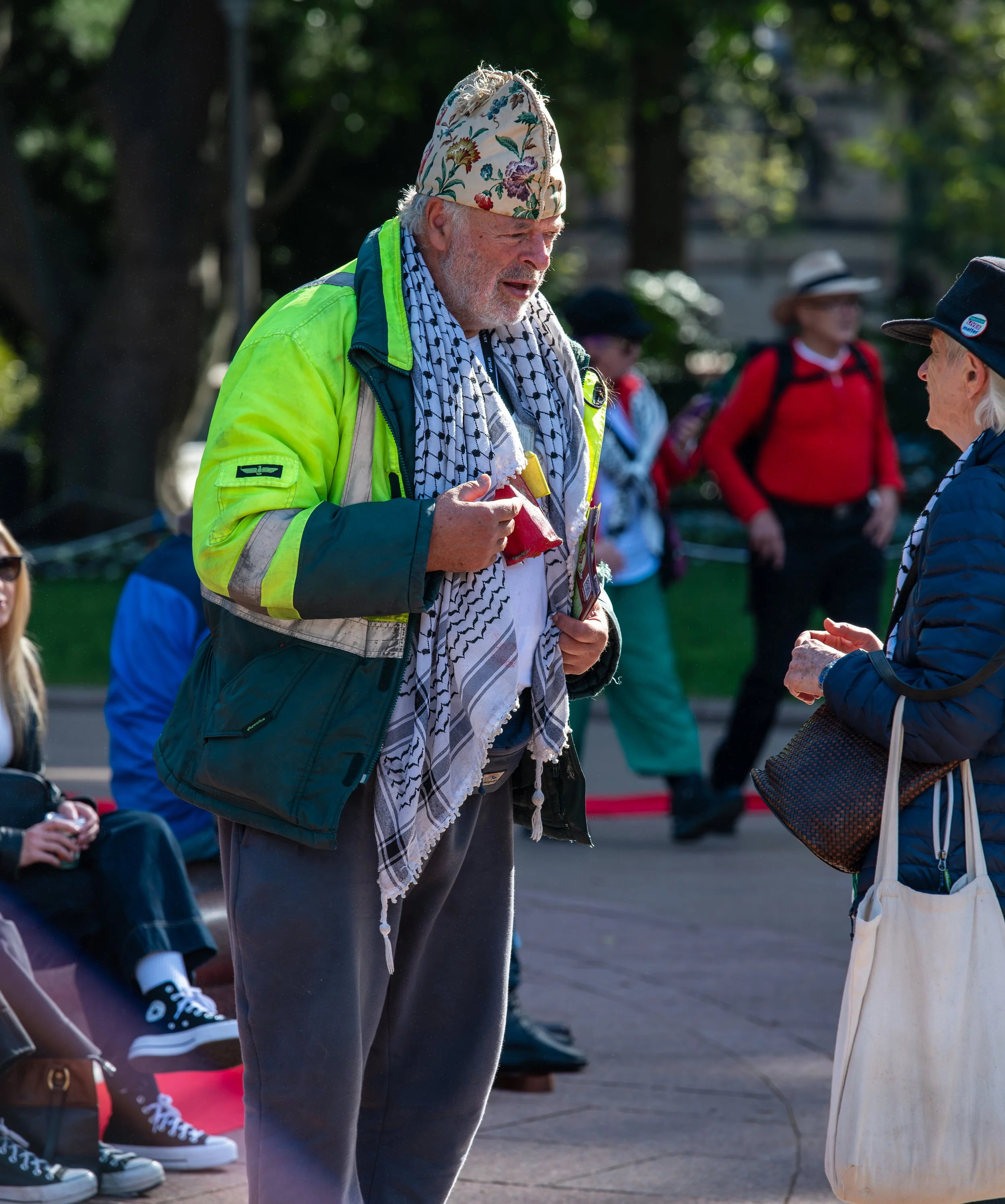 An elderly man with a white beard wearing a colorful floral hat and a yellow vest converses with a woman in a blue jacket. They are outdoors in a park with other people in the background.