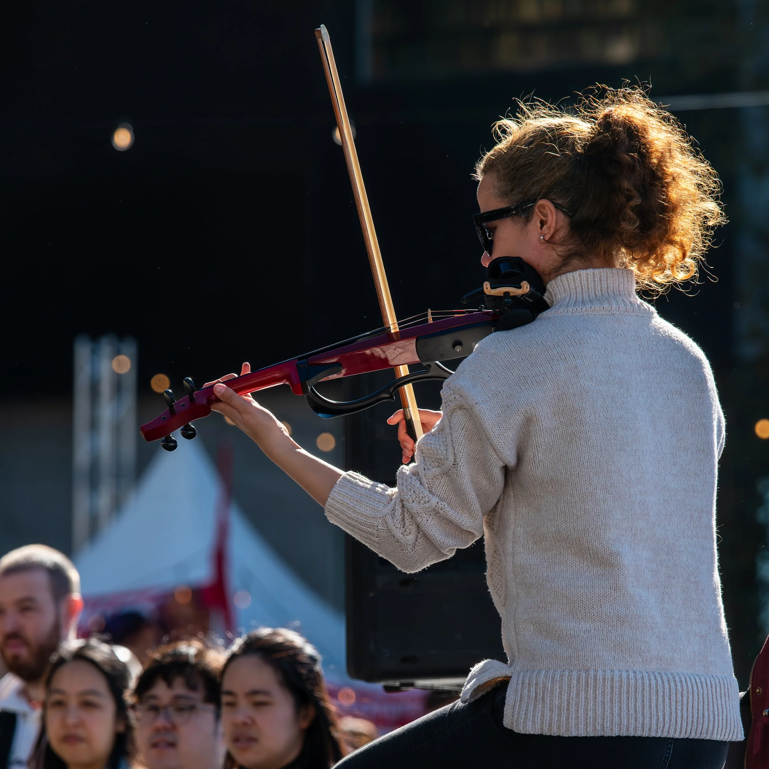 A woman with curly hair, wearing a white sweater and sunglasses, playing a violin at an outdoor event.