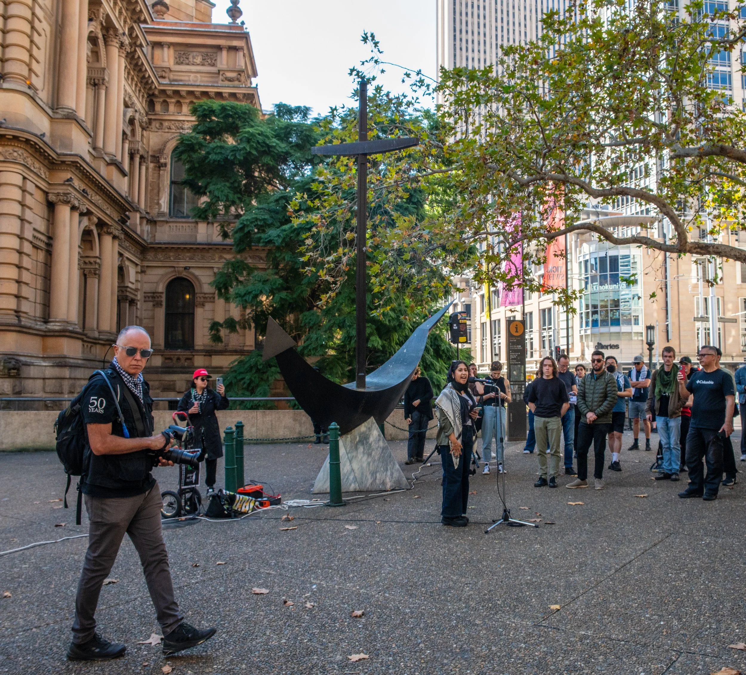 A woman speaks into a microphone in front of a modern sculpture with people gathered in an urban park setting, with historic and contemporary buildings in the background.