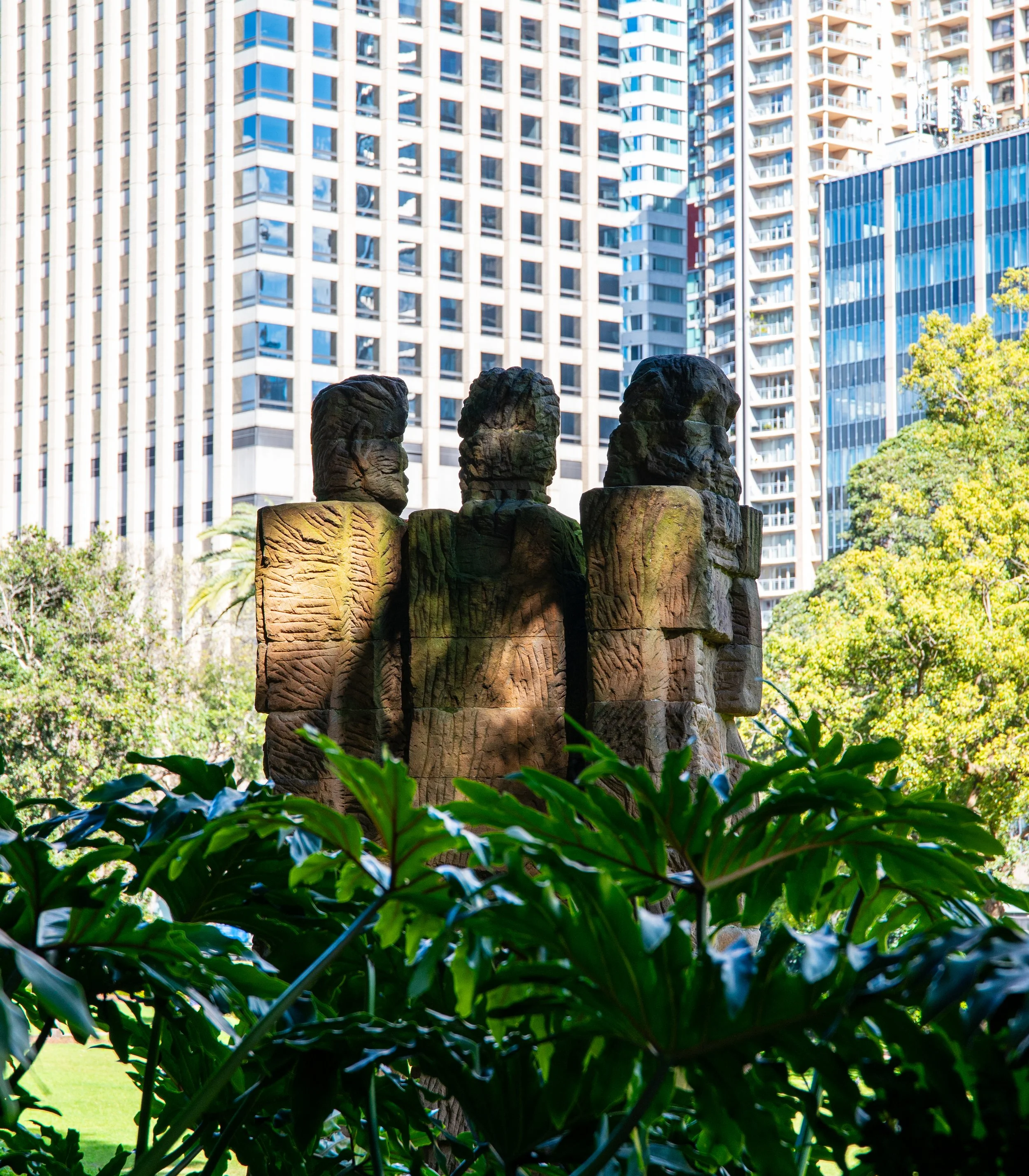 Stone sculpture of four people in front of tall modern office buildings, surrounded by green trees and plants in an urban park.