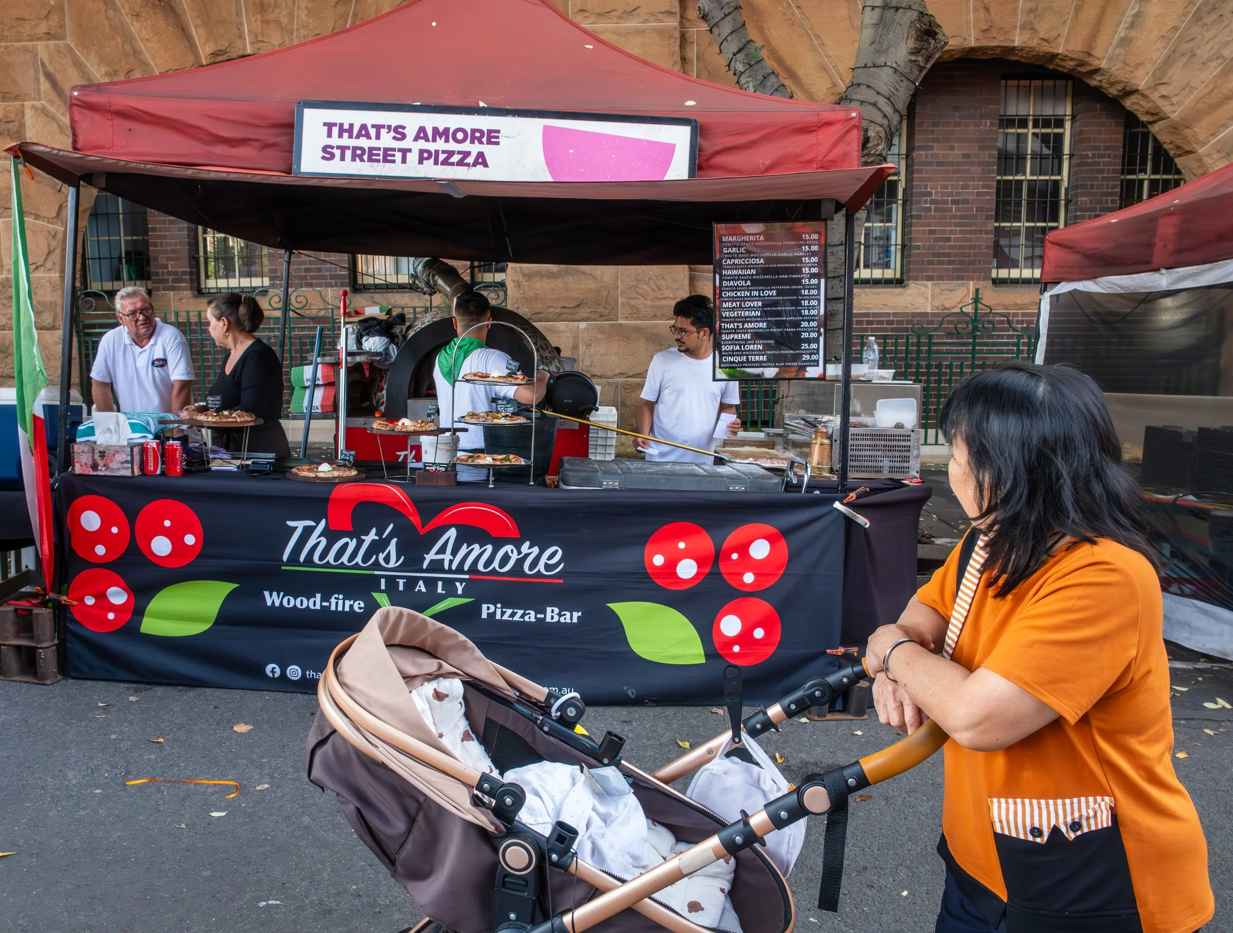 Street food stand with a pizza menu, two customers, and a woman with a stroller in front.