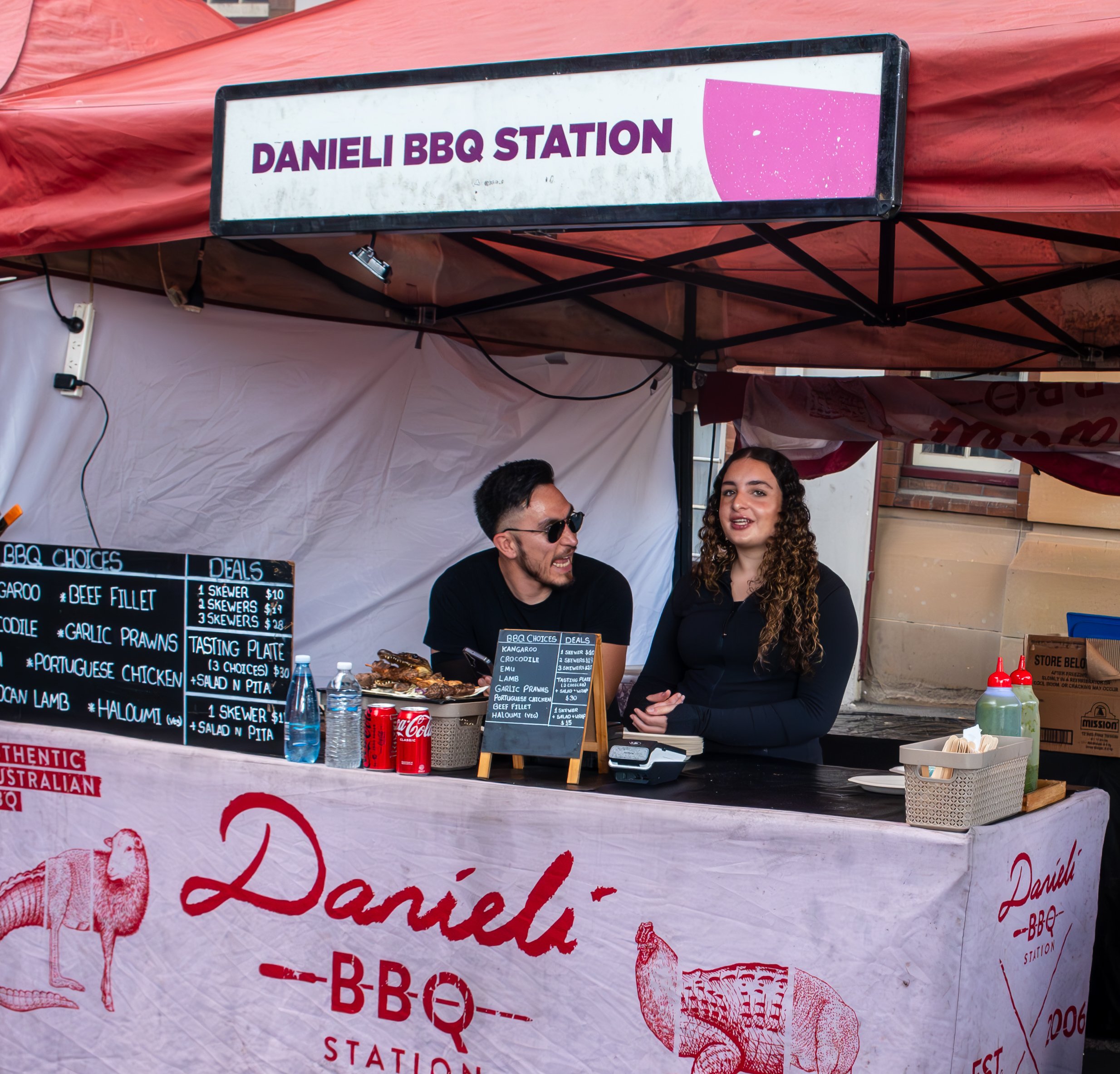 A barbecue stand with a sign reading 'Danieli BBQ Station' and two people talking behind the counter, with a menu board displaying various meat options and prices.