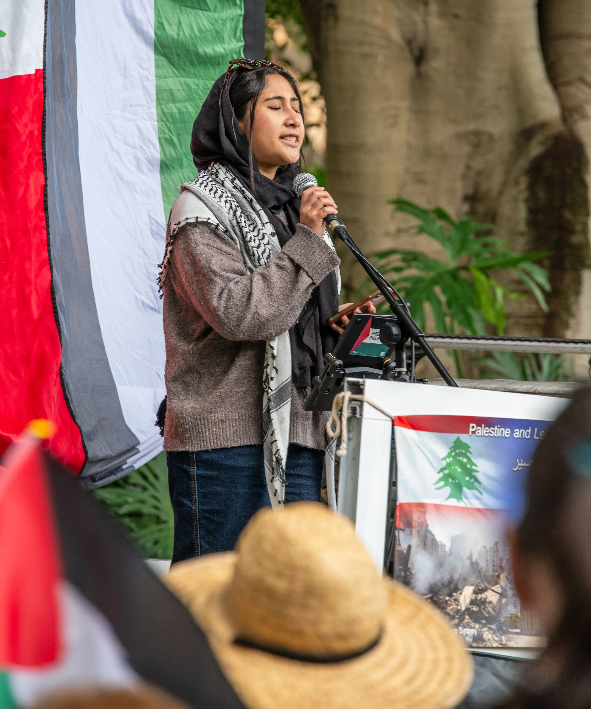 A woman speaking at a rally or protest in front of a Palestinian flag with a crowd in the foreground and a poster that mentions Palestine and Lebanon.