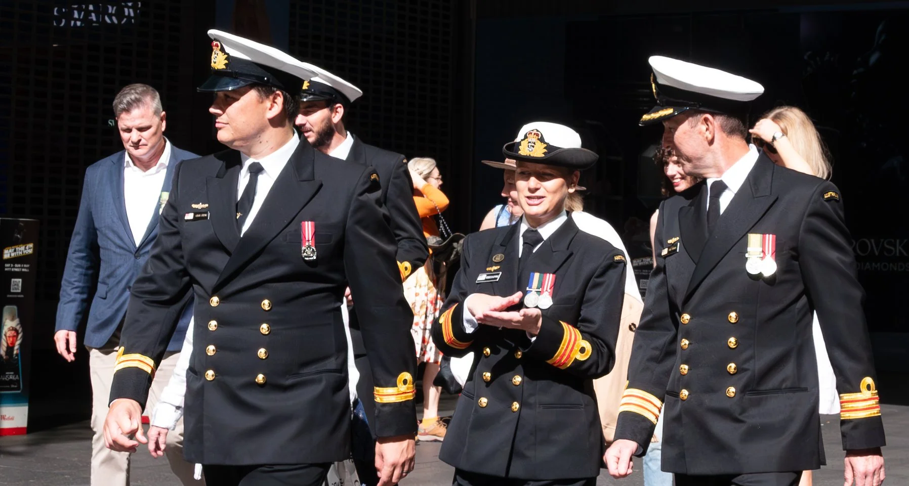 Group of Navy officers, including one woman, walking and talking on a city street during daytime. They are in formal dress uniforms with medals.