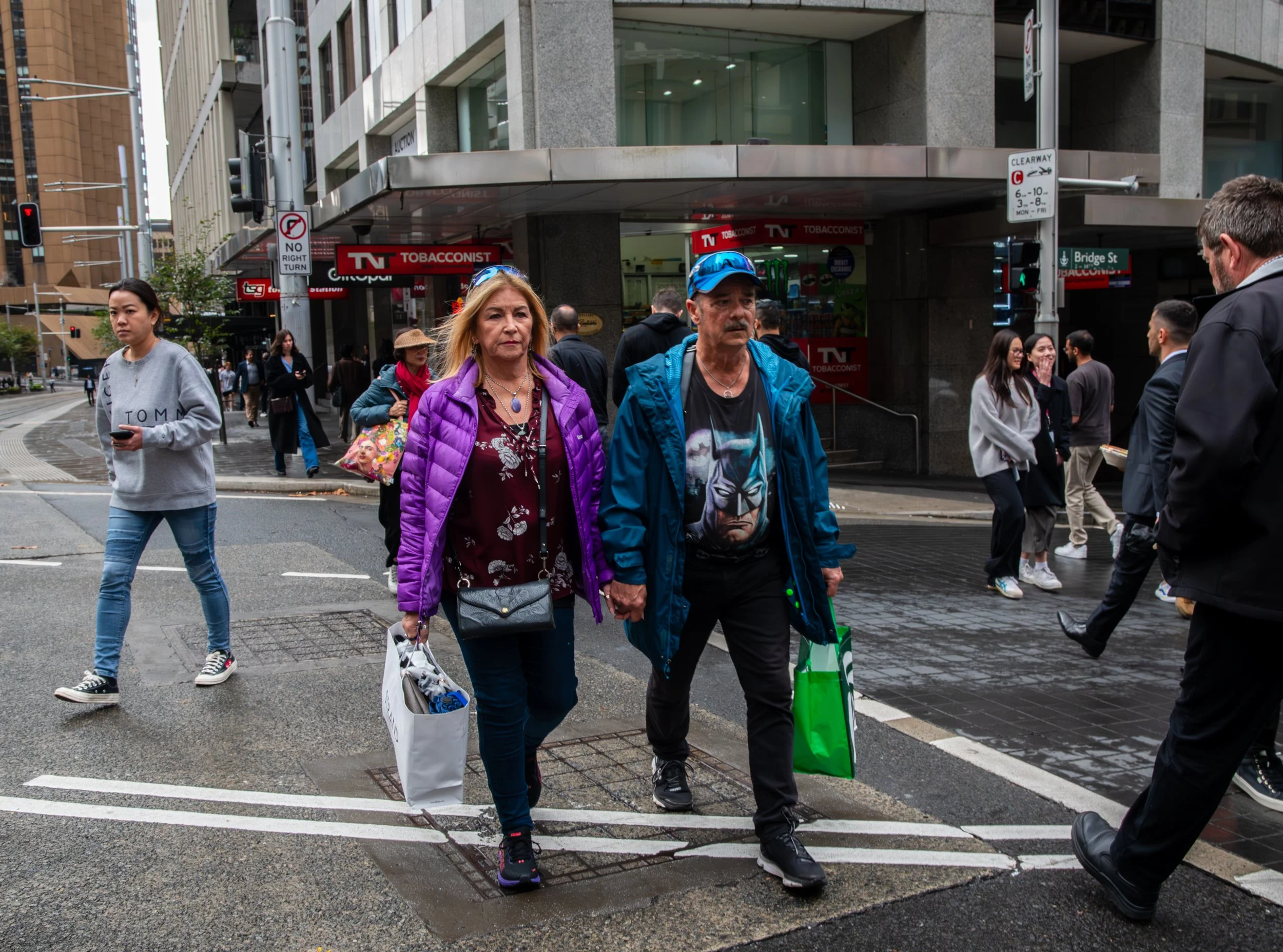People crossing a city street on a rainy day, with a building and storefronts in the background.