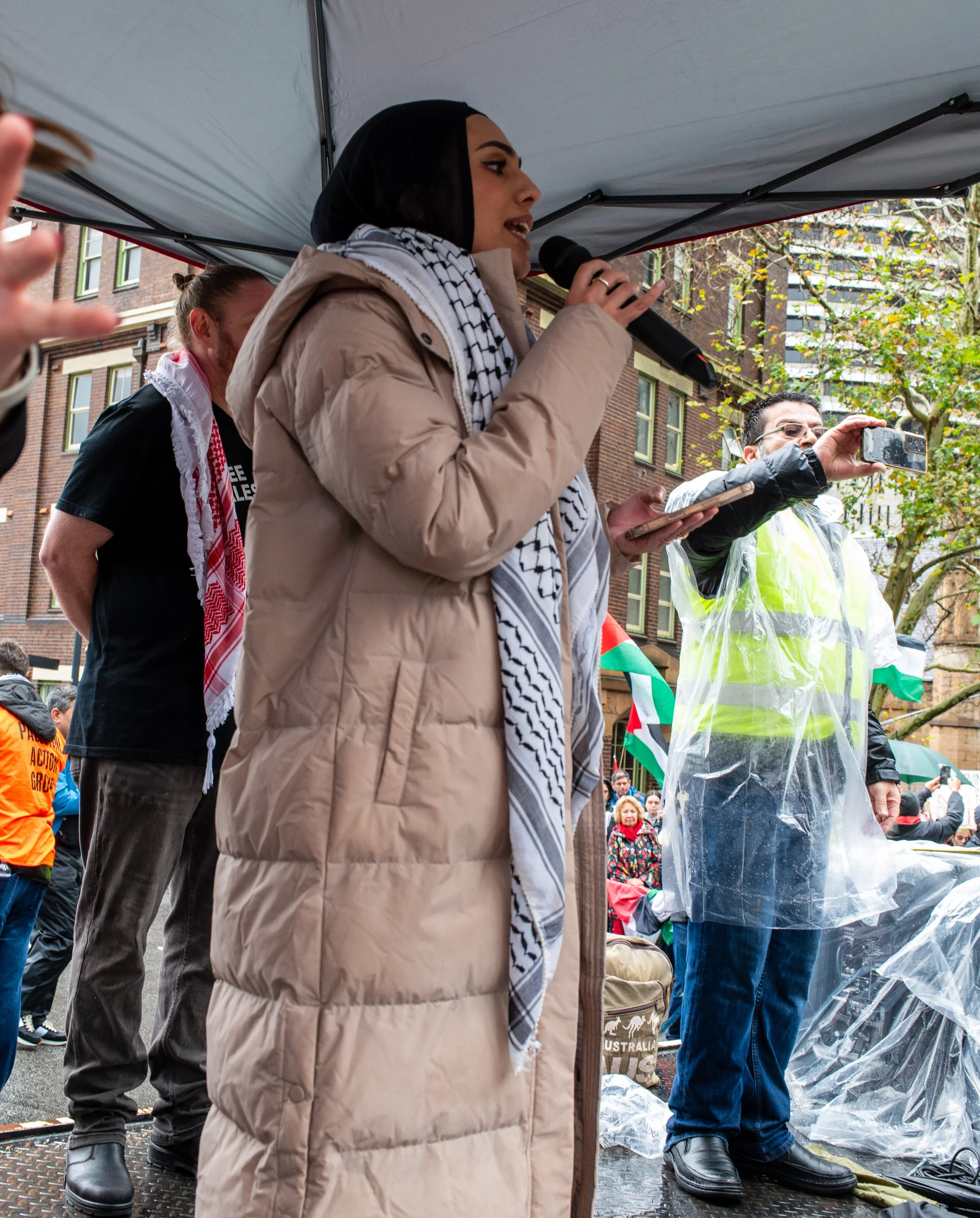 A woman speaking into a microphone at an outdoor protest or rally, with other participants and a man taking a photo or recording with a smartphone, all under a tent, with trees and buildings in the background.