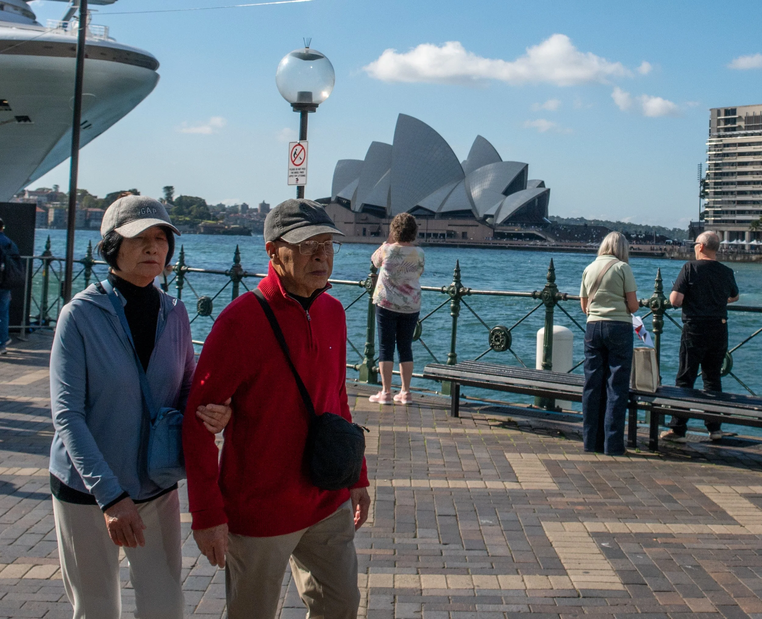 People standing on a waterfront promenade with the Sydney Opera House in the background, some looking at the water and others walking.