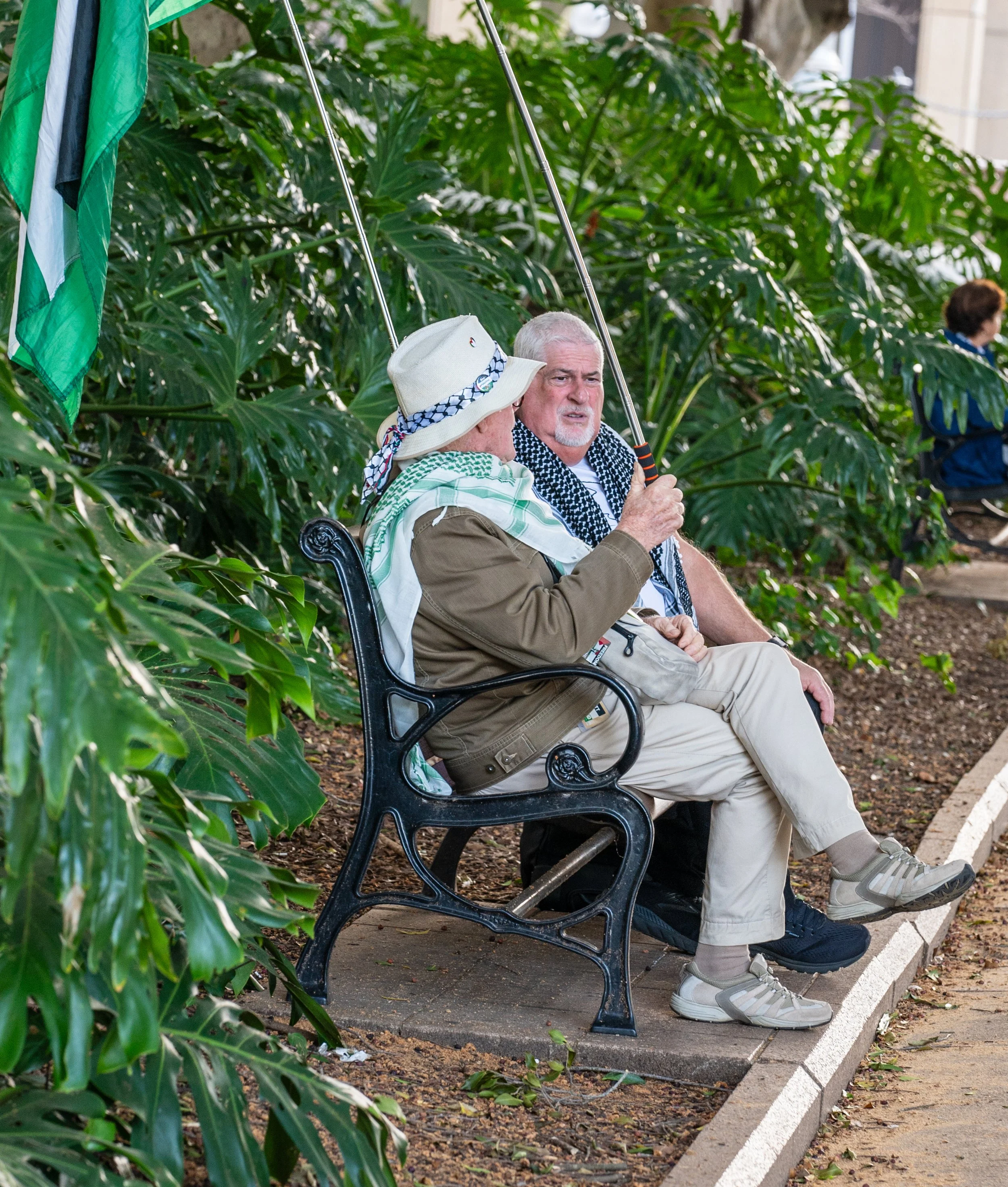 Two men sitting on a bench outside, engaged in conversation, with lush green plants in the background. One man is holding a fishing rod.