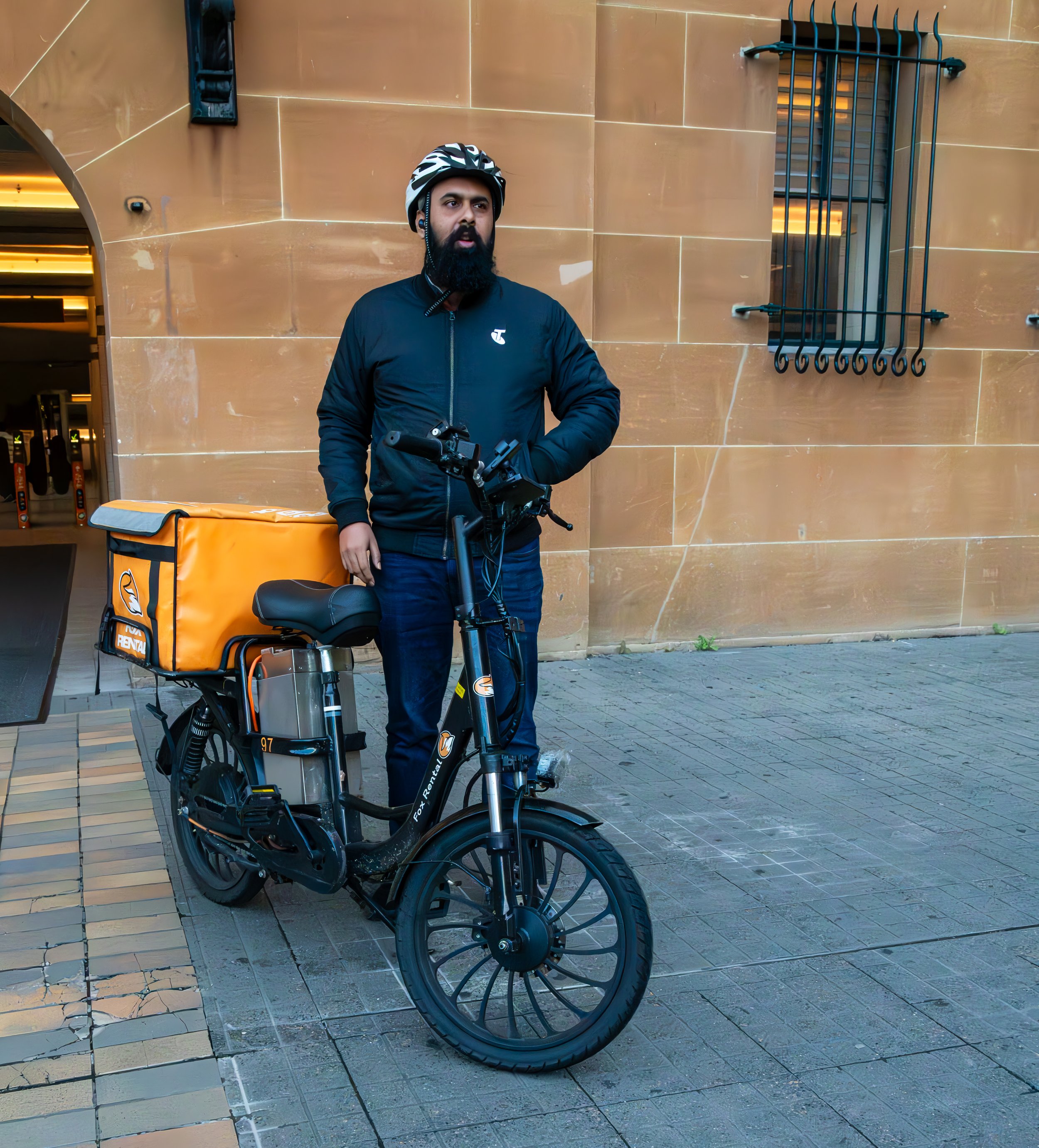 A man with a beard wearing a black jacket and a striped helmet, standing next to an orange delivery e-bike on a city sidewalk.