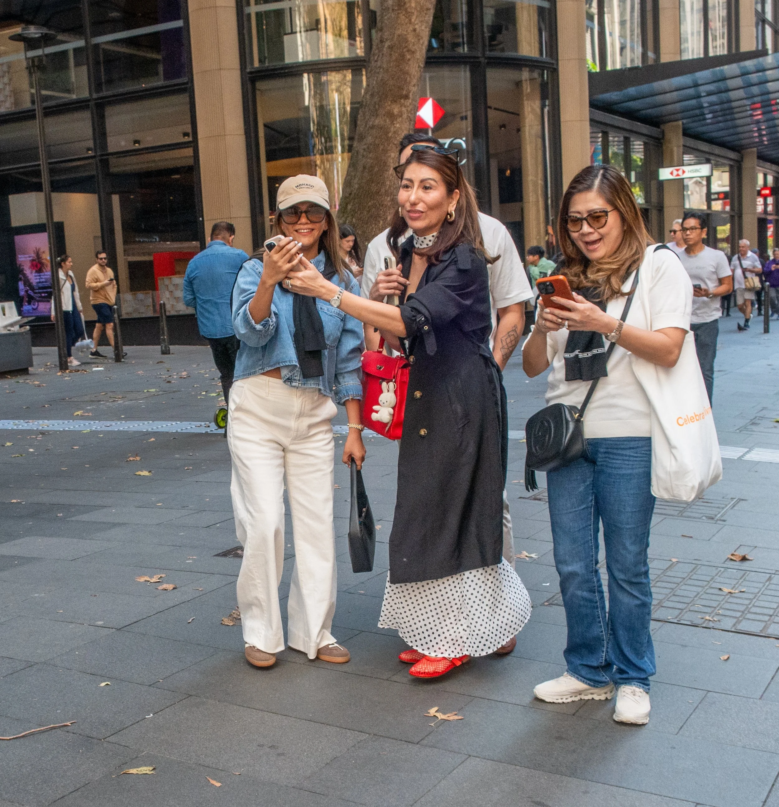 Three women standing together on a city sidewalk, looking at their phones and smiling, with other pedestrians and storefronts in the background.