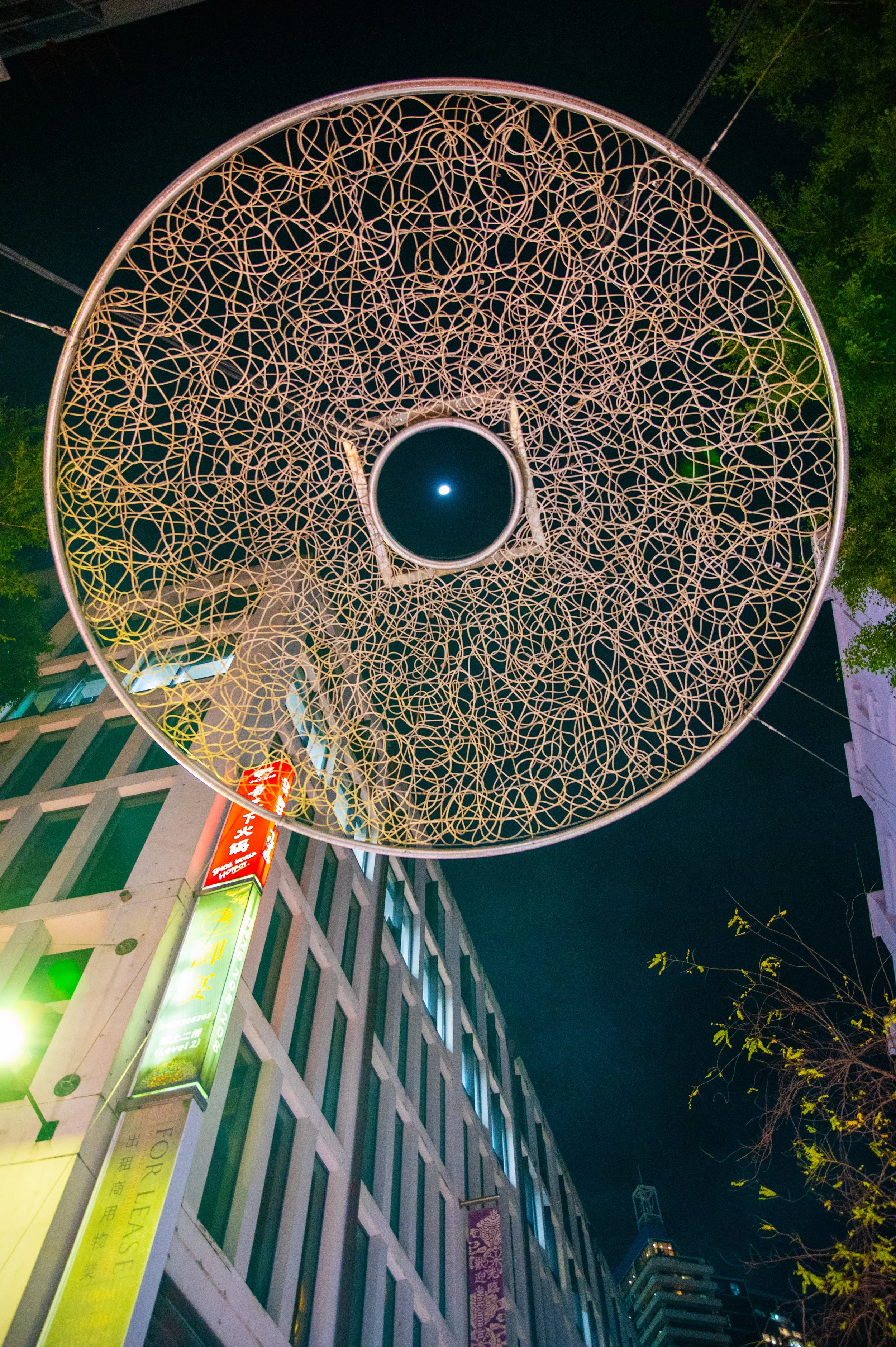 Nighttime city street view from below, featuring a large, intricate circular street decoration or sculpture with a central hole illuminated by the moon, with buildings and trees surrounding it.