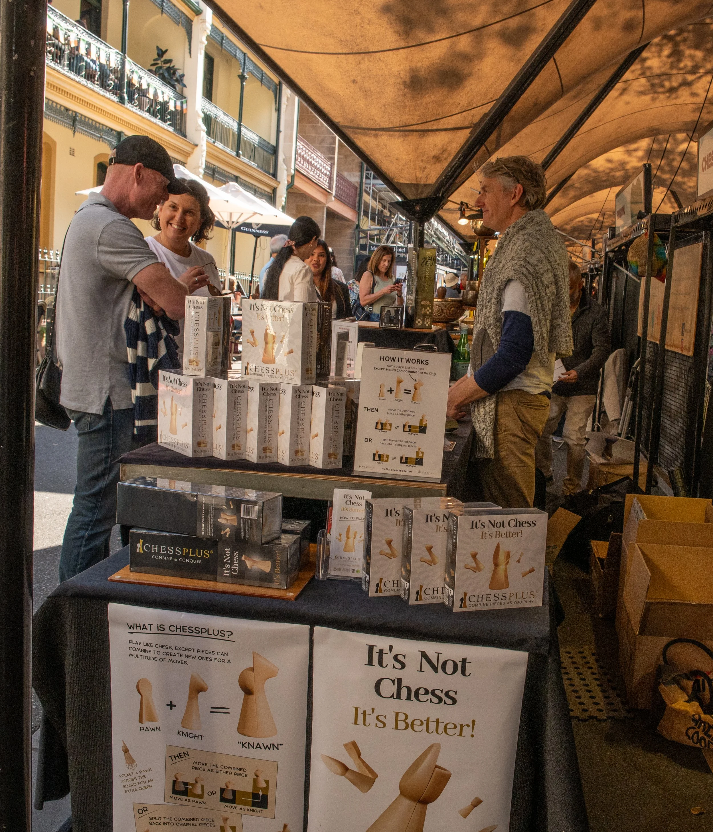 A street market booth displaying chess-inspired puzzle games called ChessPlus, with pamphlets and posters explaining how it works, as several people interact with the vendor and browse the items.