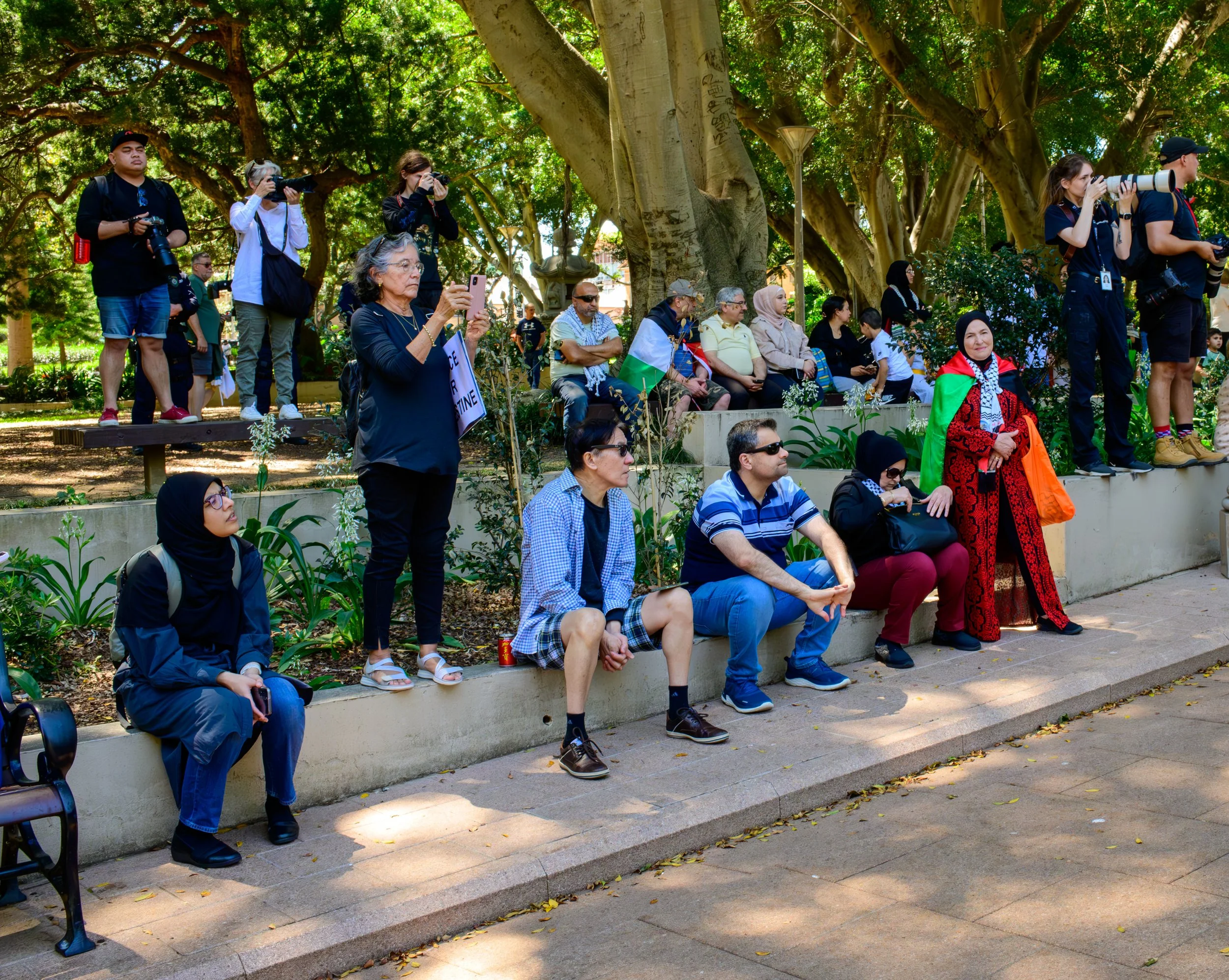 A diverse group of people gathered outdoors in a park with large trees, some sitting on benches and others standing, taking photos or watching an event, with a woman in traditional clothing and a flag visible among the crowd.