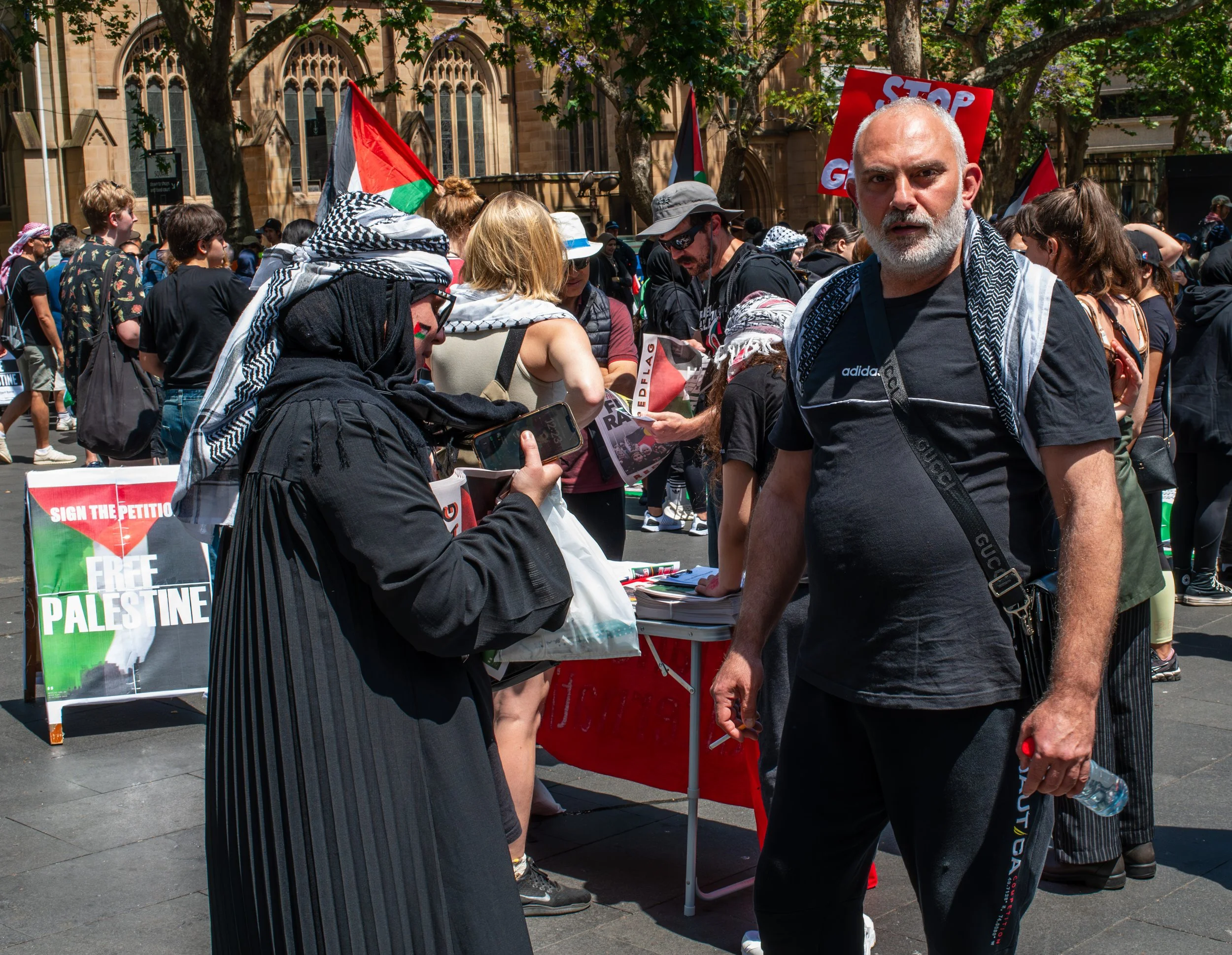 A diverse group of people at a rally or protest outside a historic building, with some holding signs supporting Palestine and Palestine flags. One sign reads 'Free Palestine.' Two individuals are engaged in conversation or activities at a table, and others are walking or standing nearby.