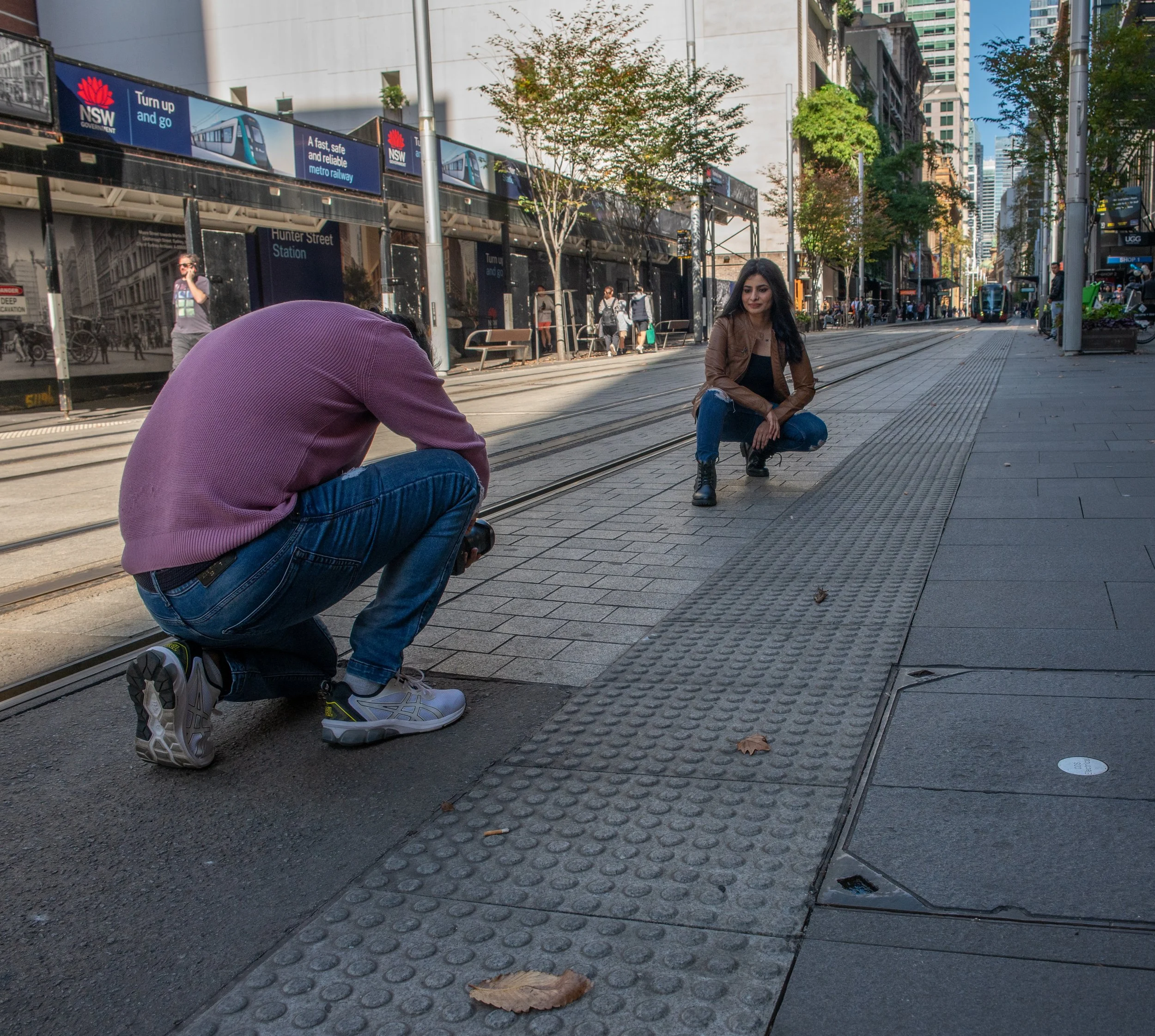 A person crouching with a camera takes a photo of a woman squatting on the sidewalk in an urban setting, with streetcar tracks, buildings, and trees in the background.