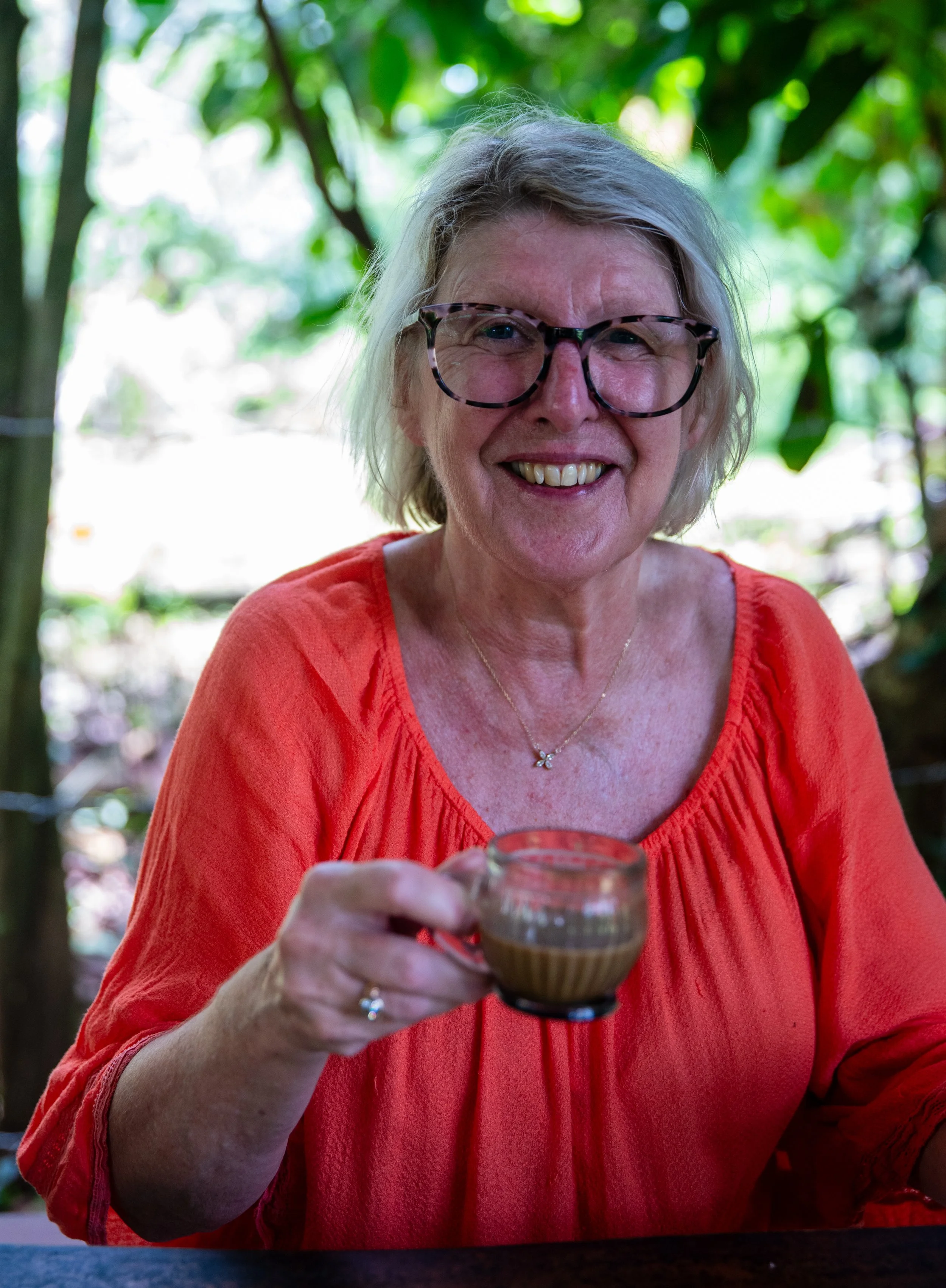 A smiling woman with gray hair and glasses holding a glass of tea or coffee outdoors with greenery in the background.