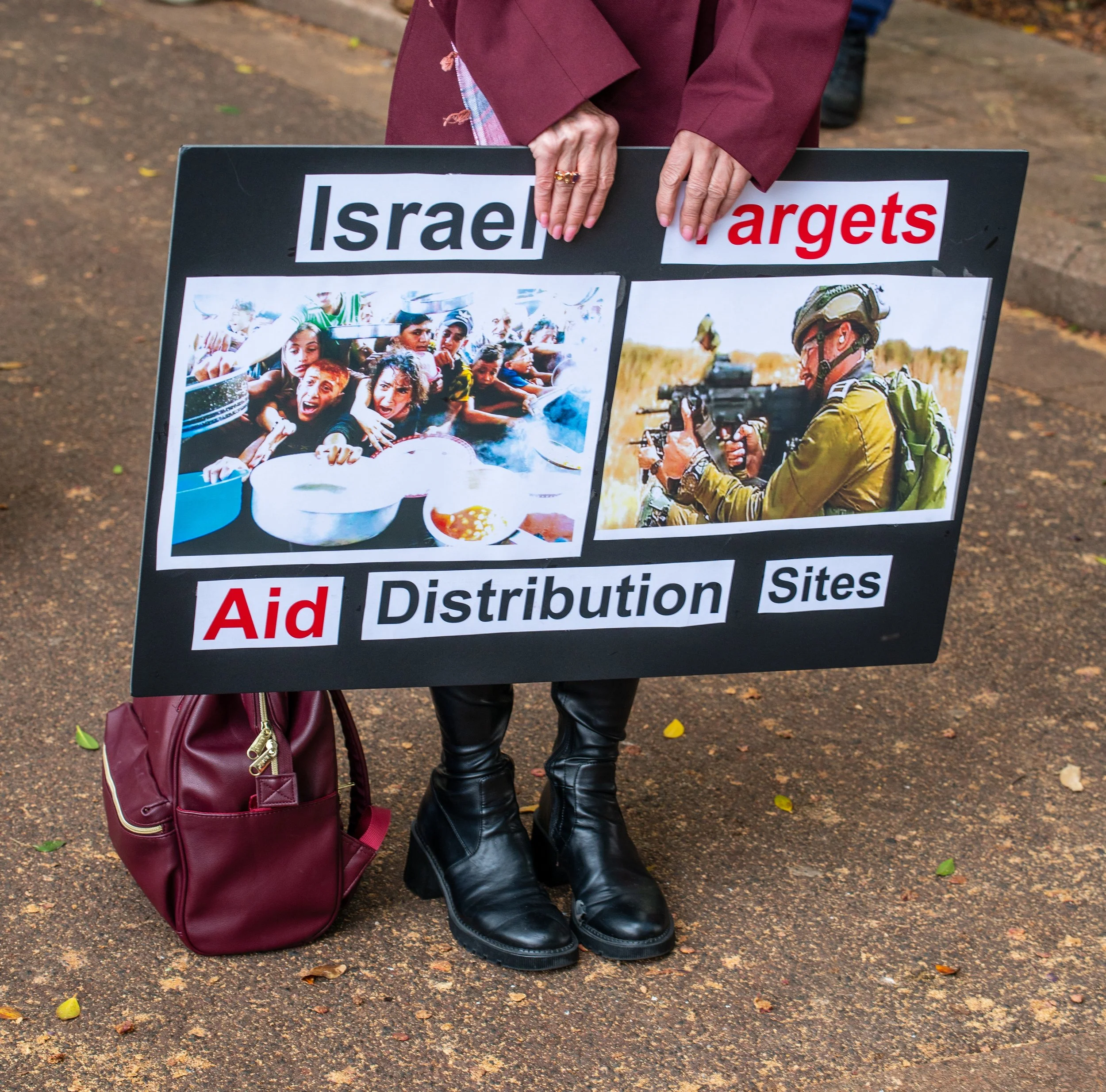 A person holding a protest sign with images and text about Israel. The sign includes a photo of children in distress and a soldier with a weapon. The words "Israel Targets" and "Aid Distribution Sites" are visible.