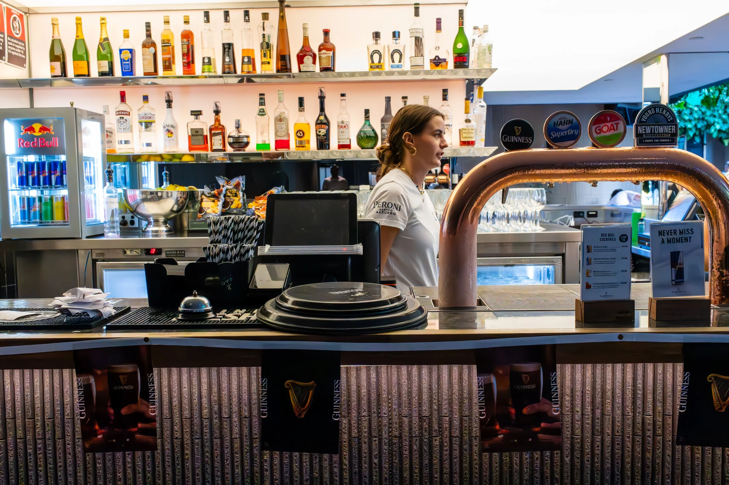 Bar scene with a female bartender behind the counter, various liquor bottles on shelves, a Red Bull fridge, and Guinness, Heineken, and other beer tap handles visible, with bar decorations and glasses ready for serving.