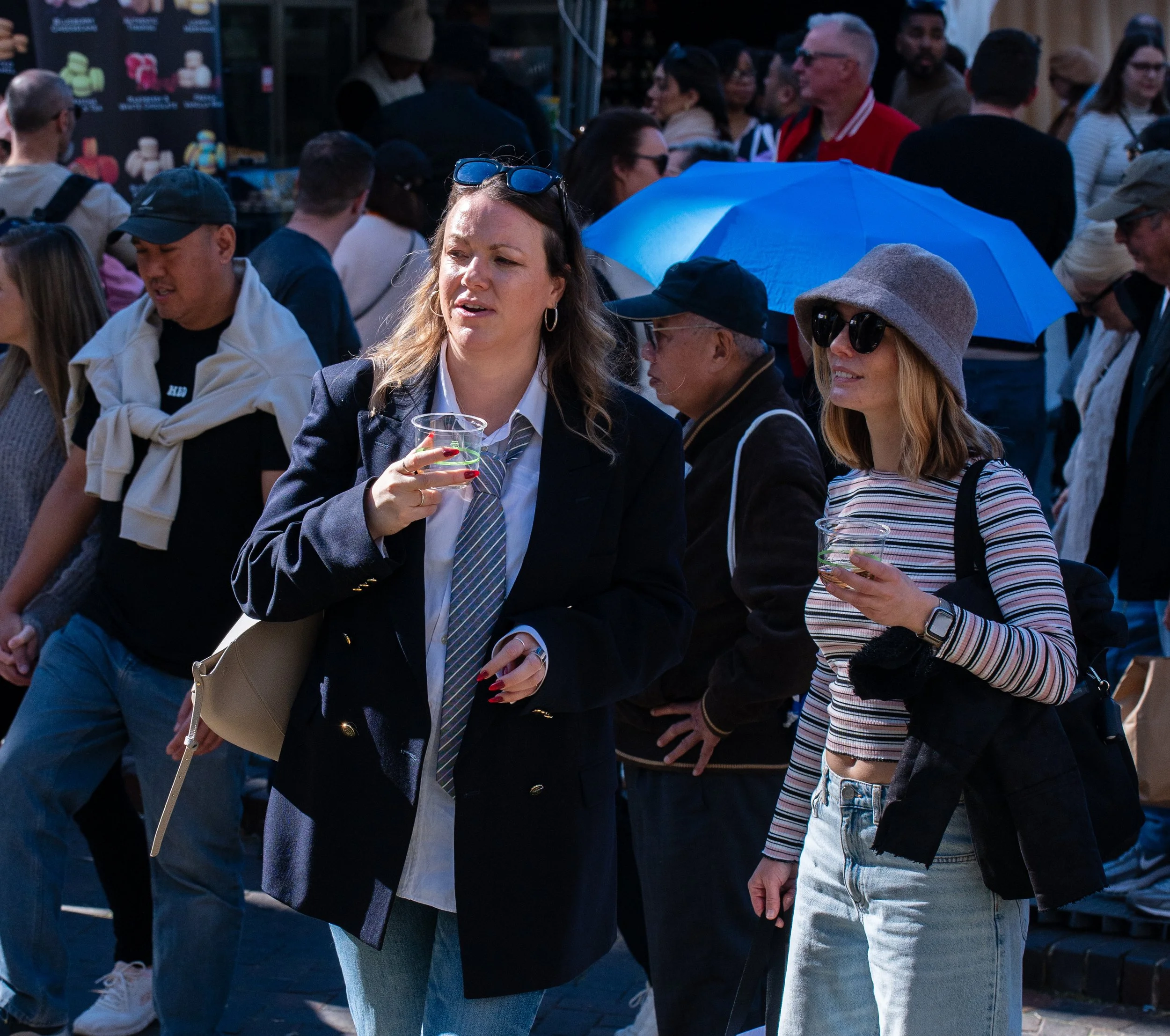 Two women standing outdoors in a crowd, holding drinks and talking. One woman wears a dark blazer, white shirt, and striped tie; the other wears a striped long-sleeve shirt, sunglasses, and a gray hat.