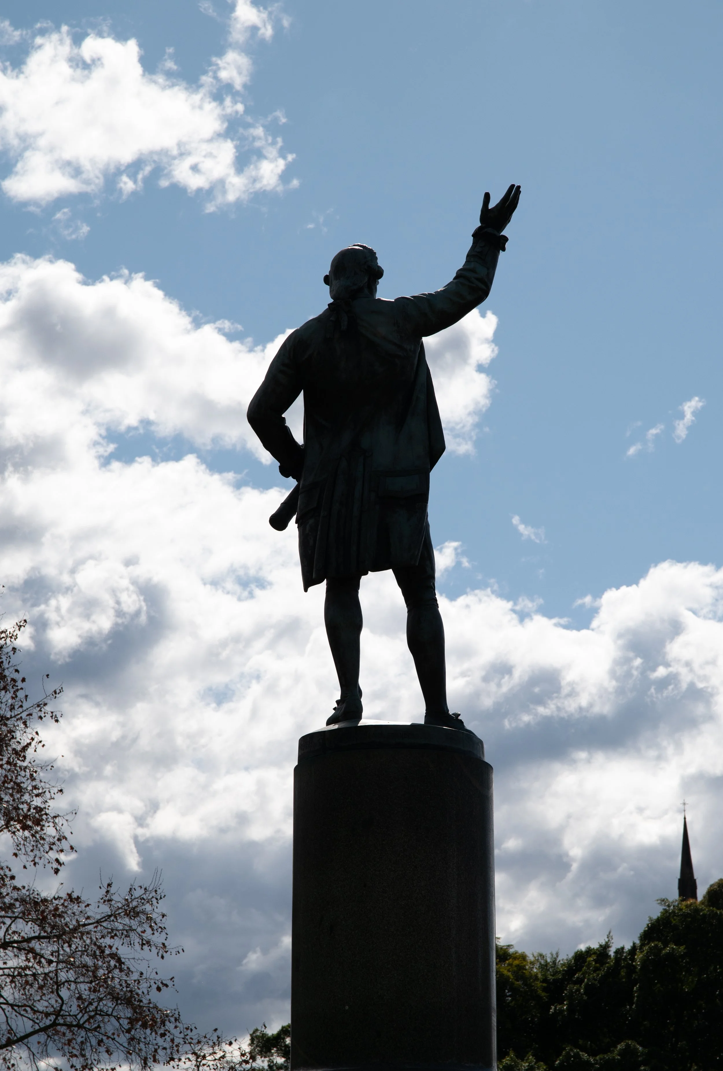 Silhouette of a statue of a man in historical attire, standing on a pedestal with his right arm raised against a backdrop of a cloudy sky.