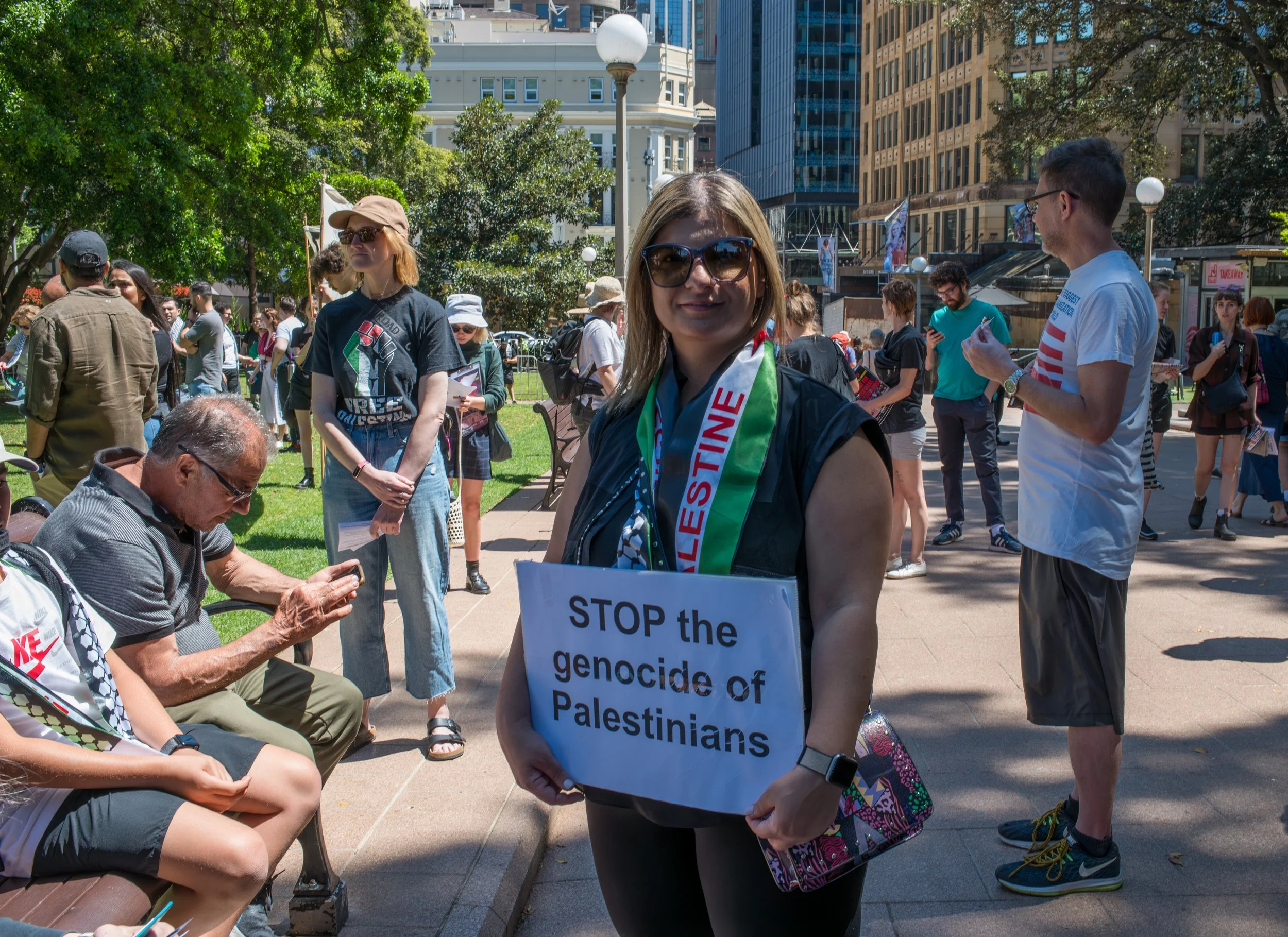 A woman wearing sunglasses and a scarf stands in a crowd holding a sign that reads, 'STOP the genocide of Palestinians.' People around her are standing or sitting, engaging in conversations or on their phones, in a city park during daytime.