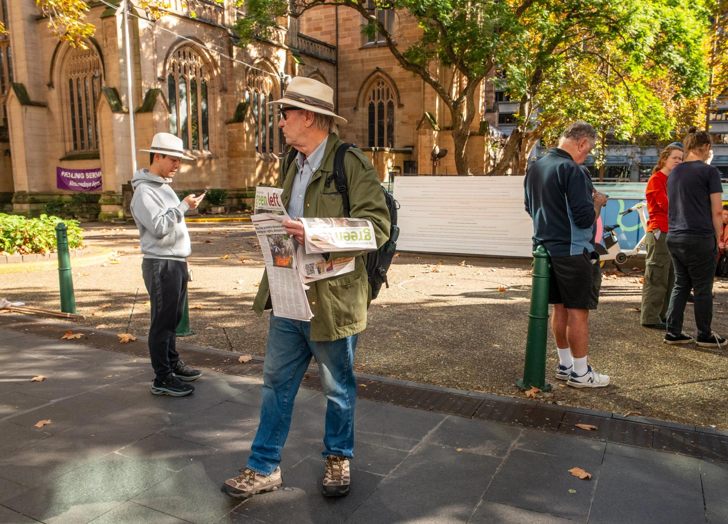 People standing in line outdoors near a historic building, with one man reading a newspaper.