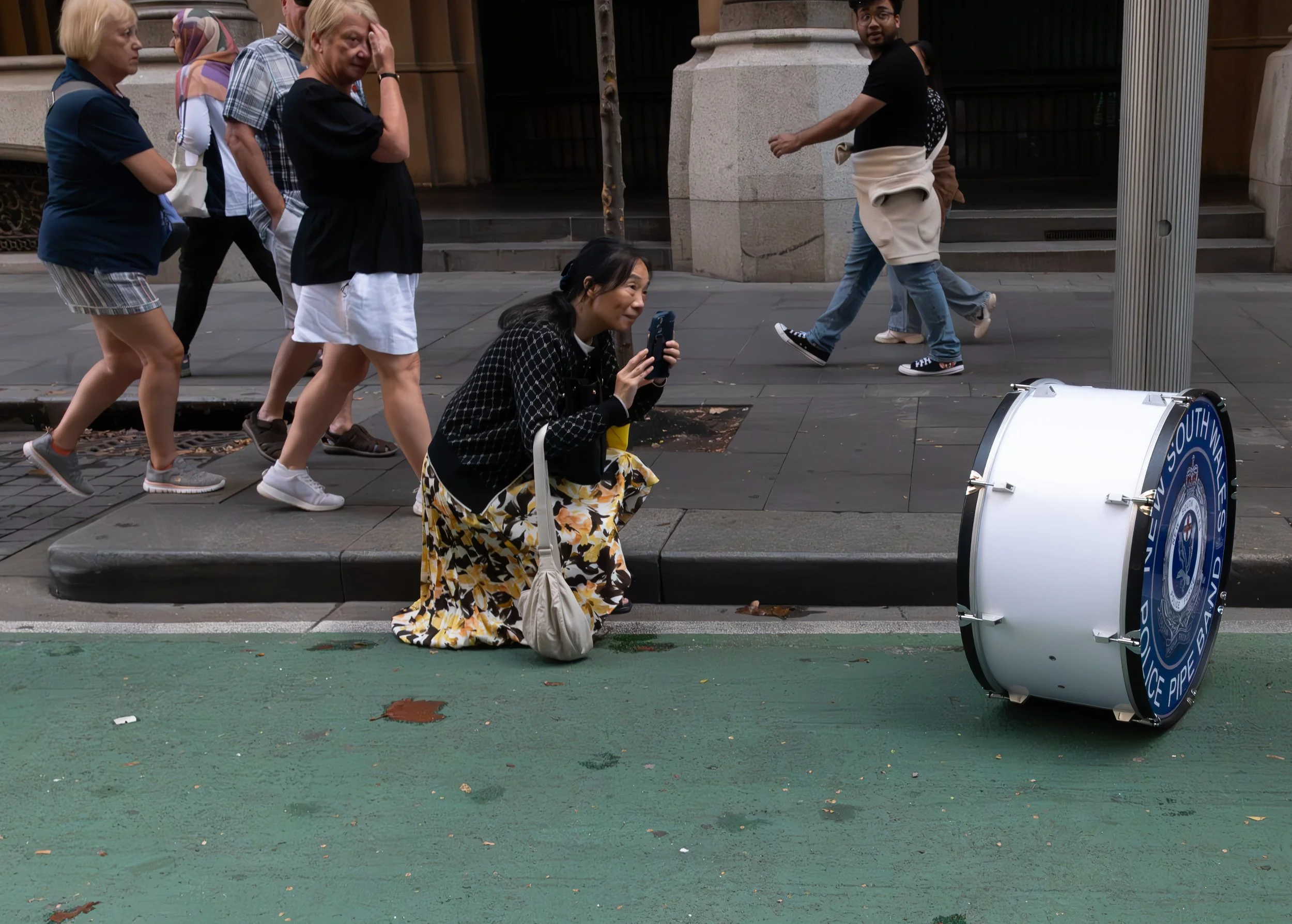 A woman crouching on the sidewalk using her phone to take a picture of a large police drum on a green bike lane, with pedestrians walking behind her on a city street.