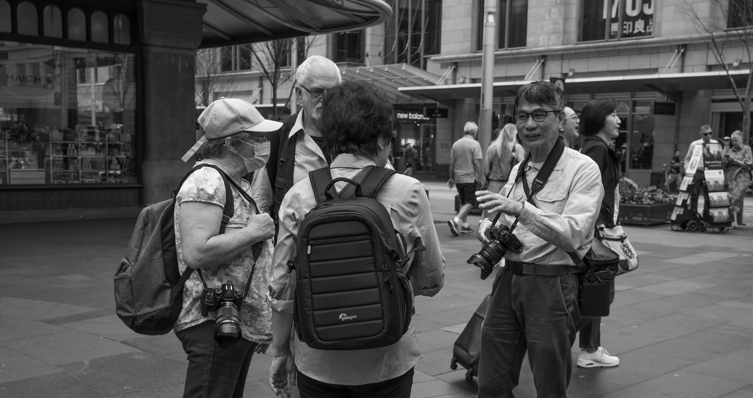 A group of tourists with cameras and backpacks talking on a city street, possibly in a shopping or downtown area.