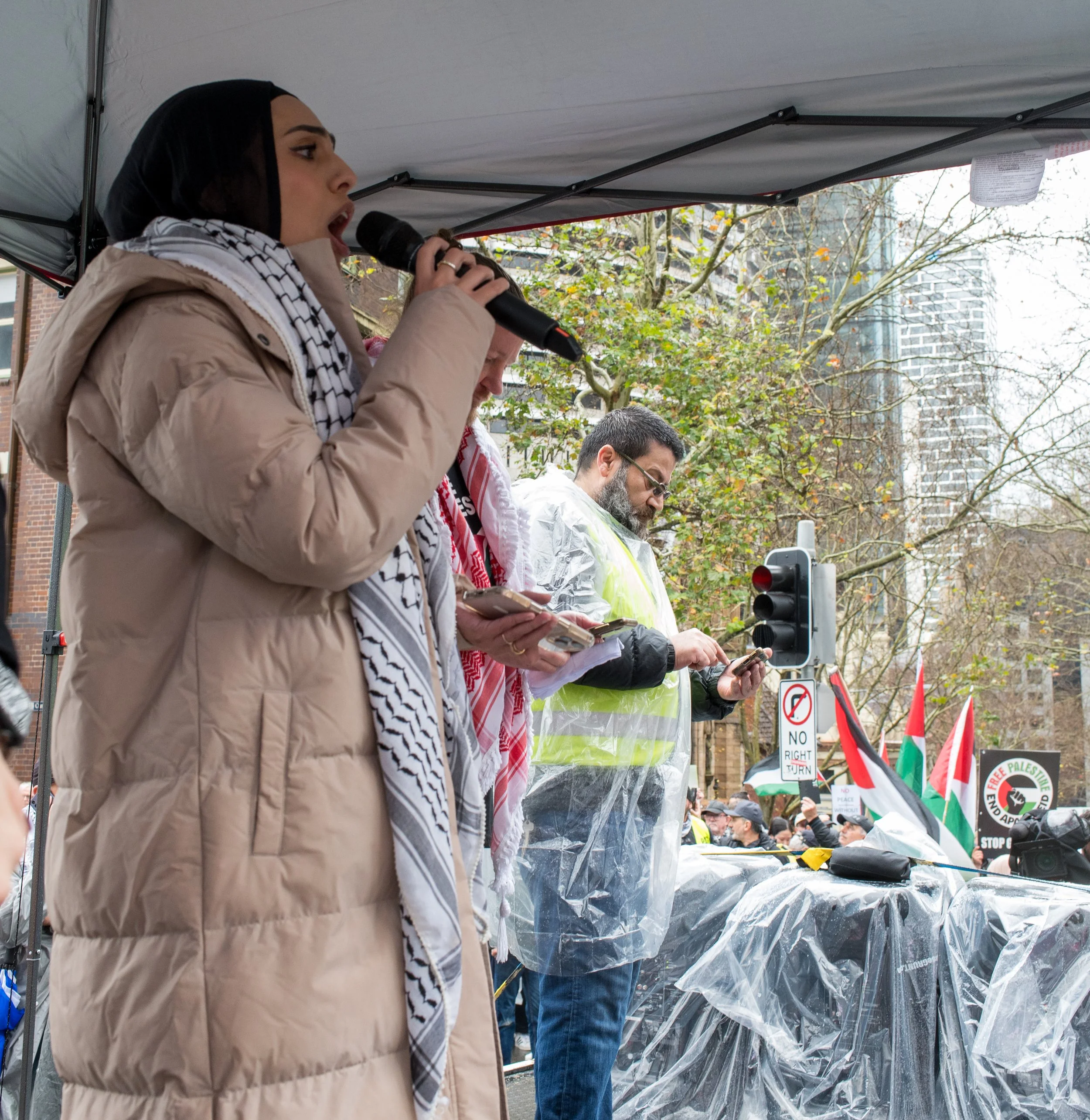 People speaking and using phones at a protest or rally, with flags and signs in the background.