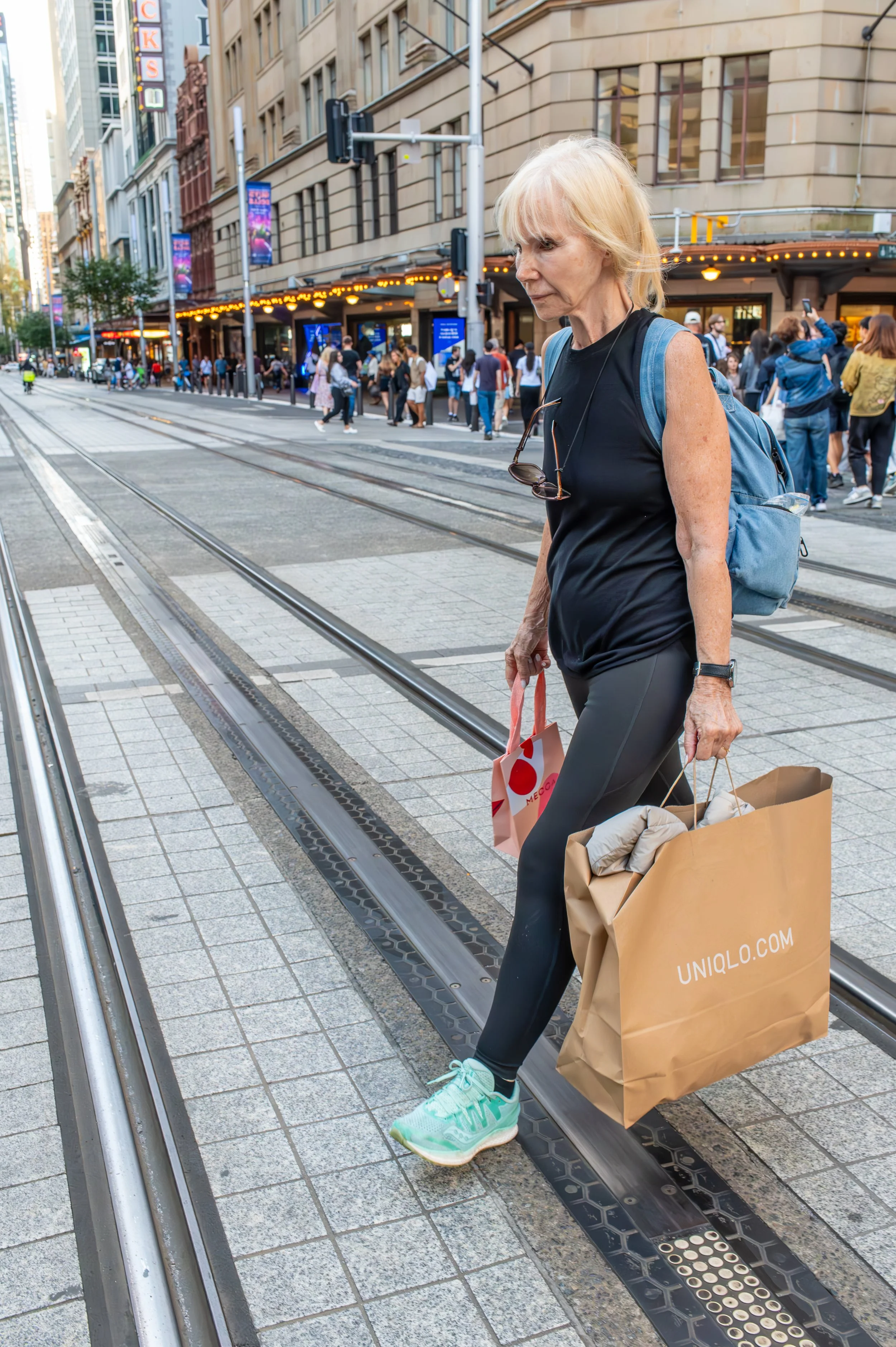 An elderly woman crossing a city street with tram tracks, carrying shopping bags and wearing athletic clothing and sneakers.