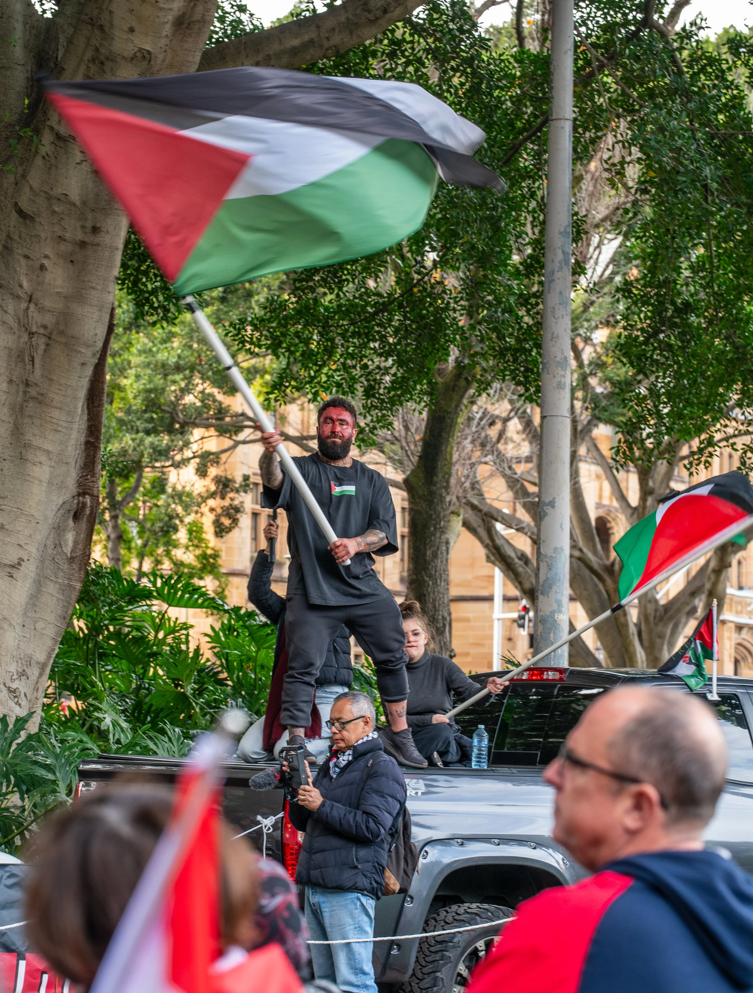 A man with a beard and tattoos stands on the back of a truck, waving a Palestinian flag during a demonstration. Others around him are holding Palestinian flags, and there are trees and a historic-looking building in the background.