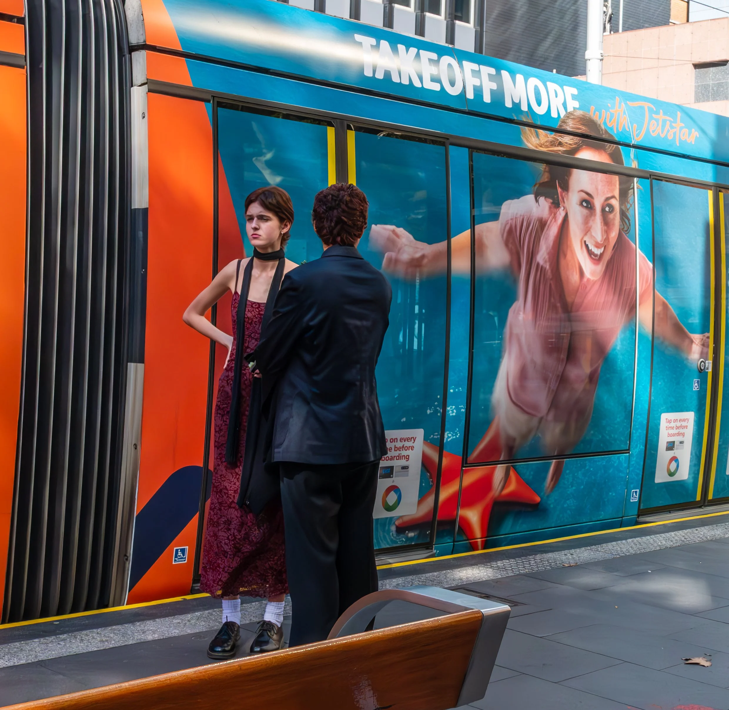Two people standing in front of a colorful advertisement on a city transit station. The ad features a woman swimming underwater and the text 'Takeoff More with Jetstar'.