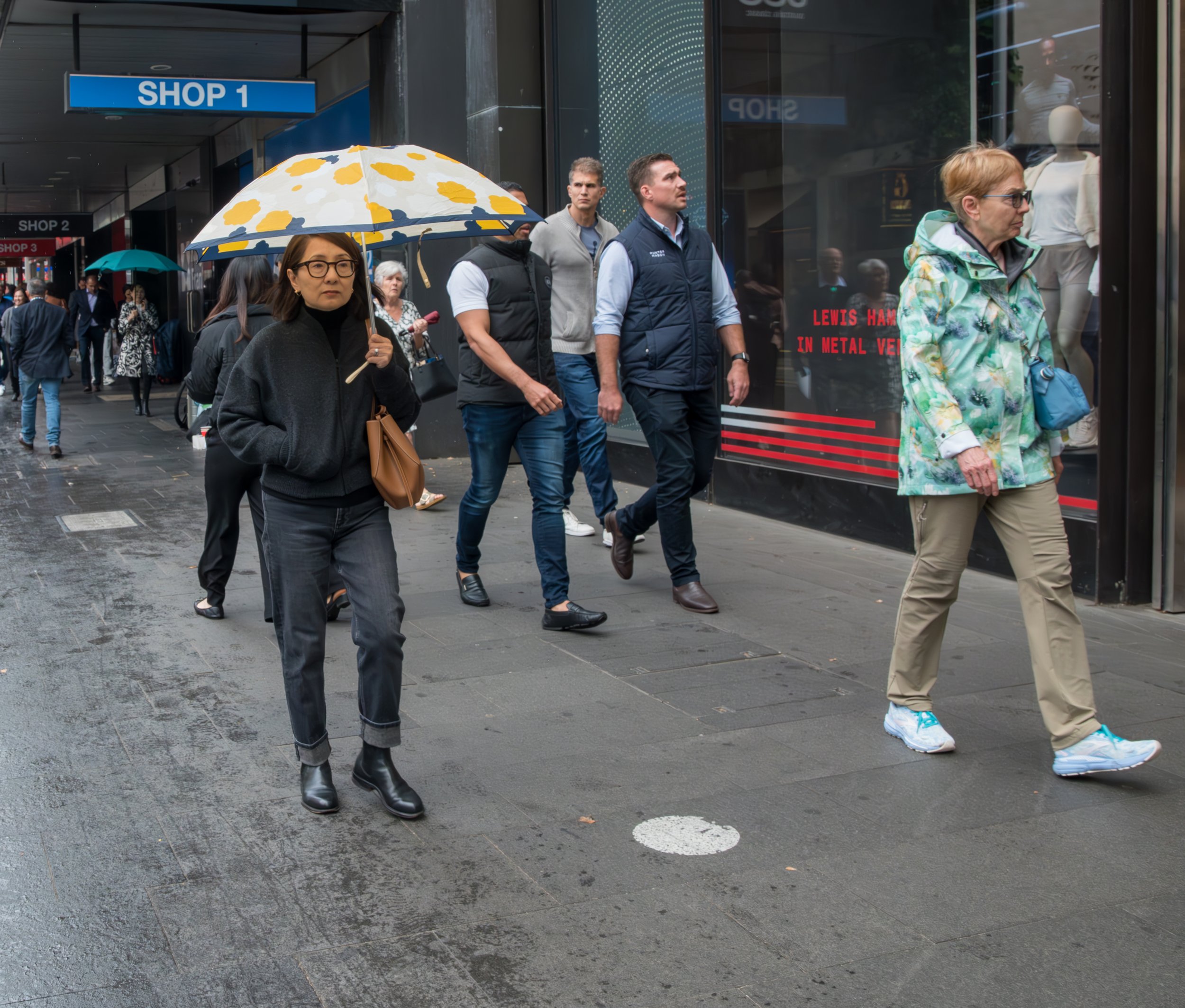 People walking on a rainy city sidewalk, some with umbrellas, in front of shops with large windows and signage.